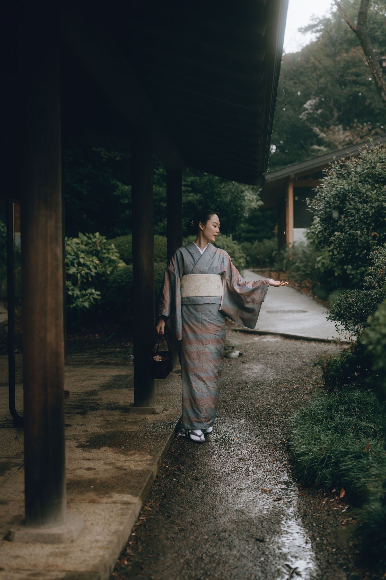 Woman in traditional gray kimono with obi belt walking through a serene Japanese garden pathway.
