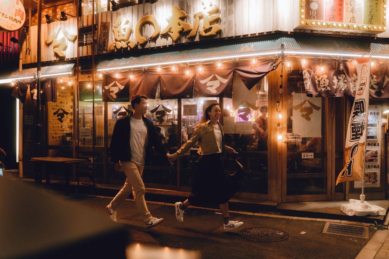 Couple walking past a warmly lit Japanese restaurant at night with glowing lanterns and neon signs in yurakucho, tokyo.