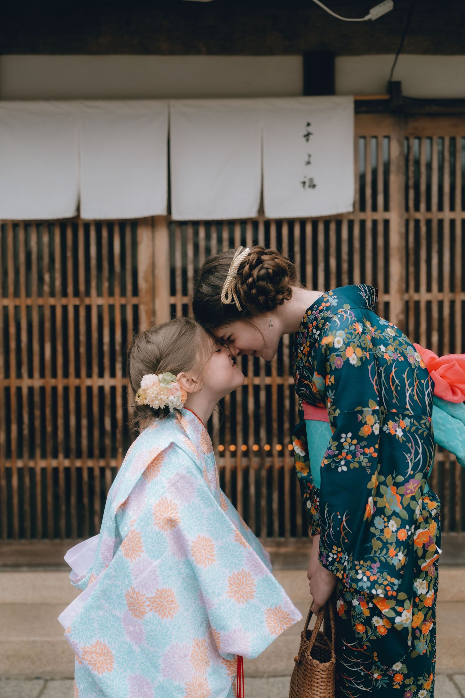A mother and daughter in matching traditional Japanese kimonos share a tender forehead touch outside a bamboo building.Sawara, Chiba, Japan.