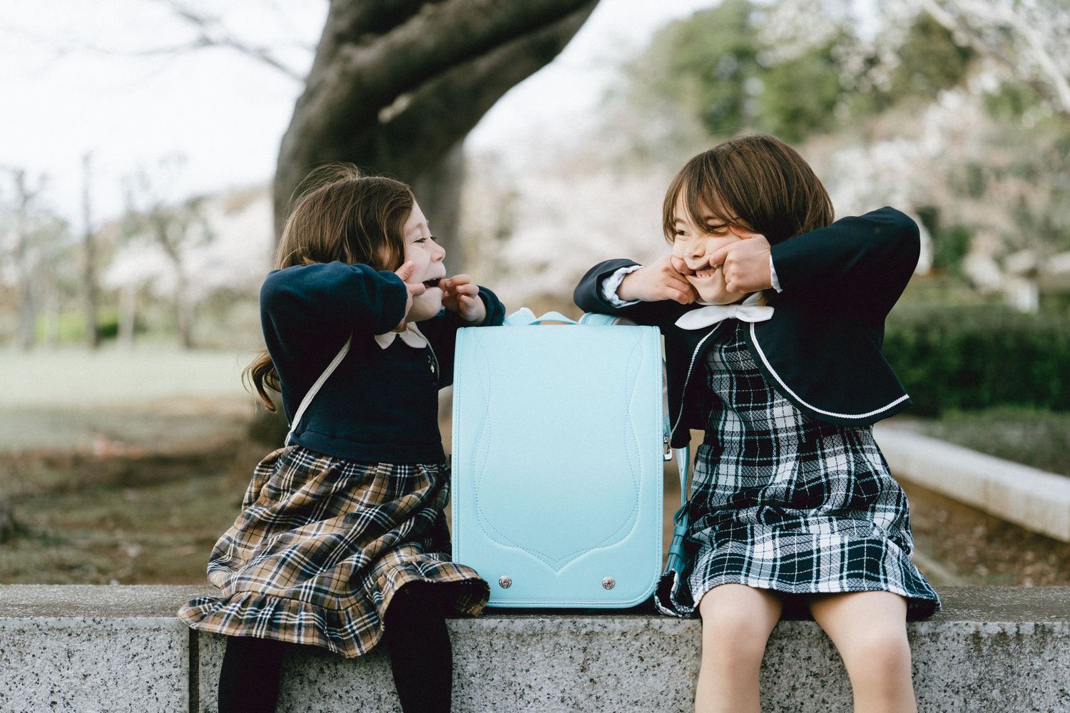 Two young girls in school uniforms sit on a stone bench, laughing and gesturing over a light blue backpack outdoors.