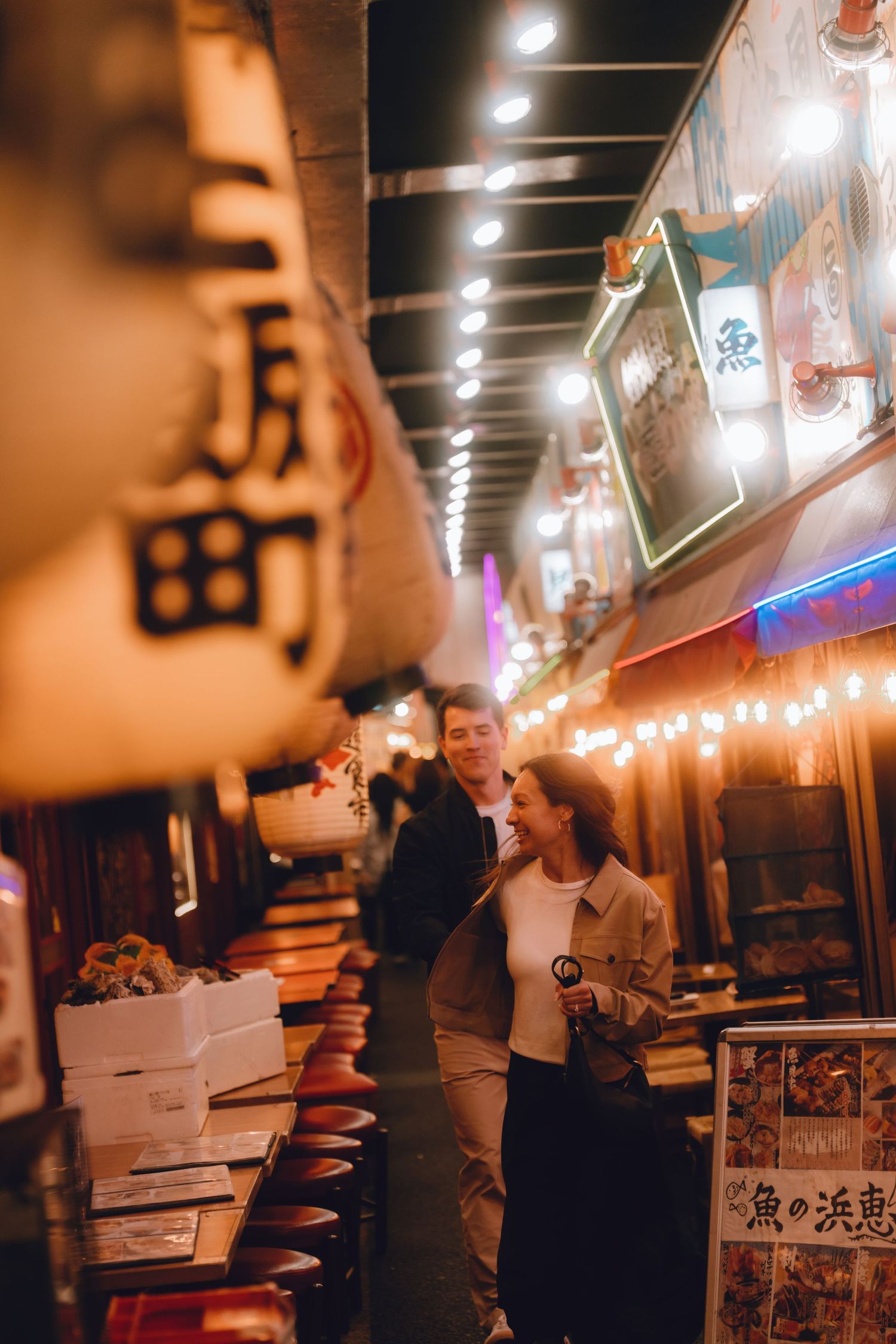 Couple smiling together at a vibrant Japanese night market with glowing lanterns and neon signs in yurakucho, tokyo.