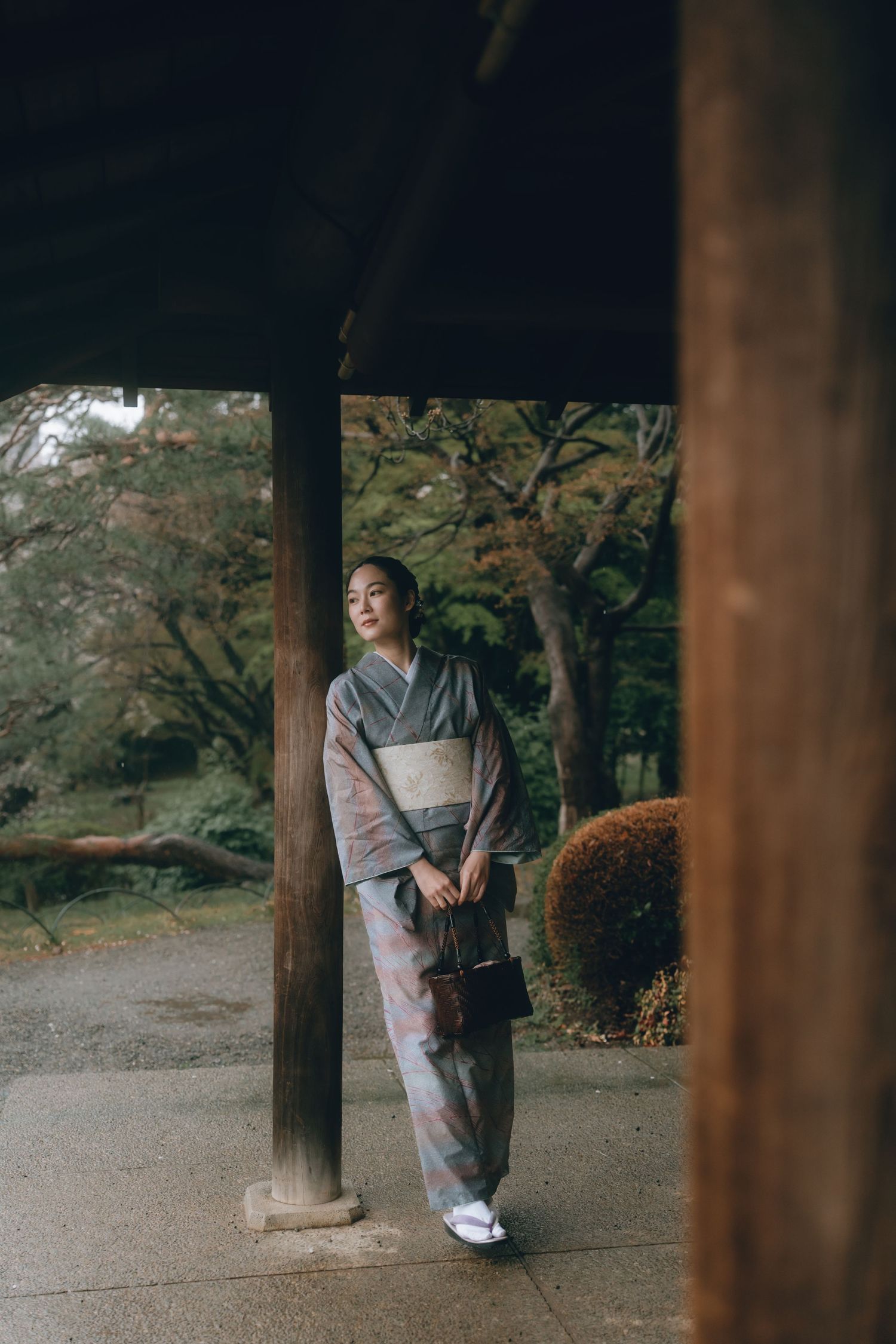 A woman in a traditional gray Japanese kimono with obi walks through a serene garden path beside wooden pillars  in Shinjyuku gyoen, Tokyo,