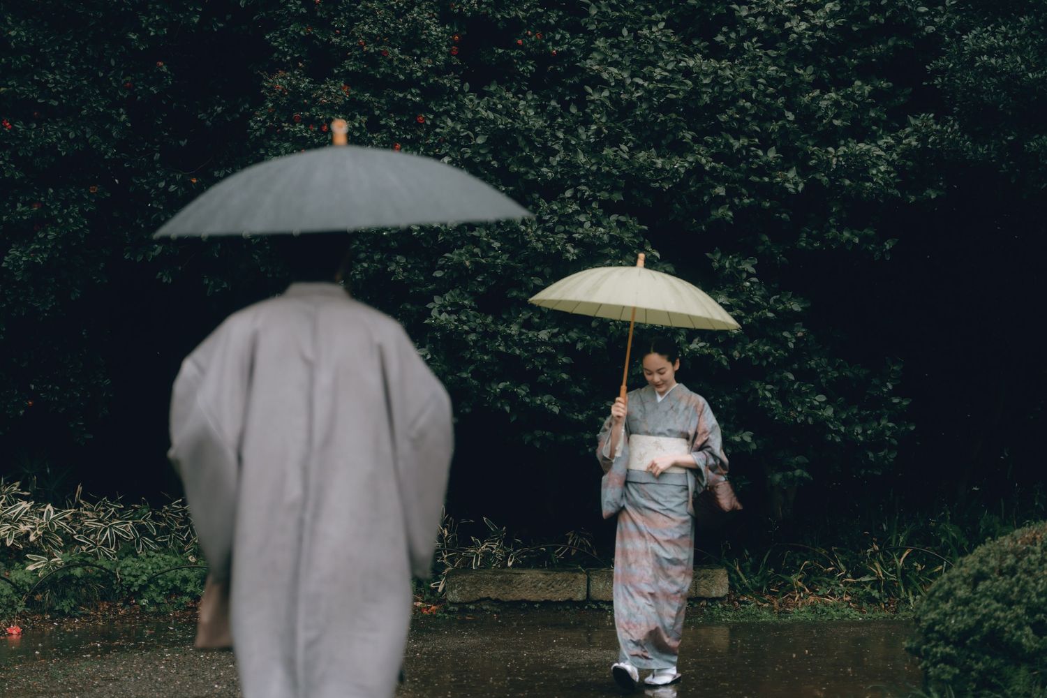 Two women in traditional Japanese kimonos walk through a lush garden in the rain, each holding a delicate umbrella in Shinjyuku gyoen, Tokyo