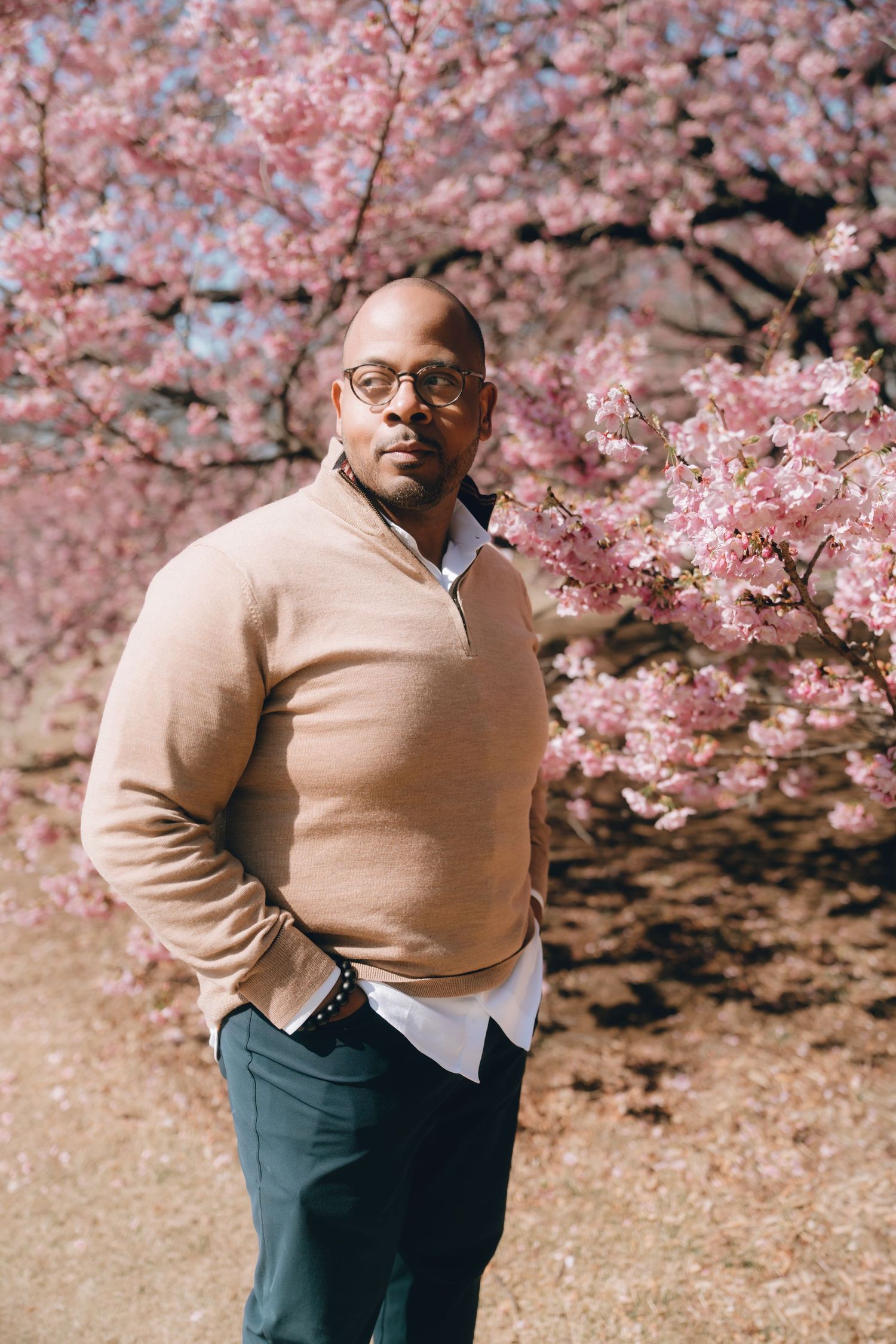 A man in glasses and beige sweater stands confidently before blooming pink cherry blossom trees.