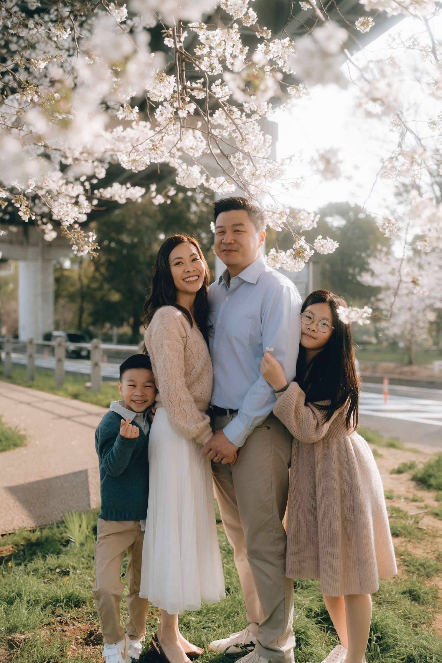Happy Asian family of four posing under blooming cherry blossom trees in spring, smiling together outdoors.