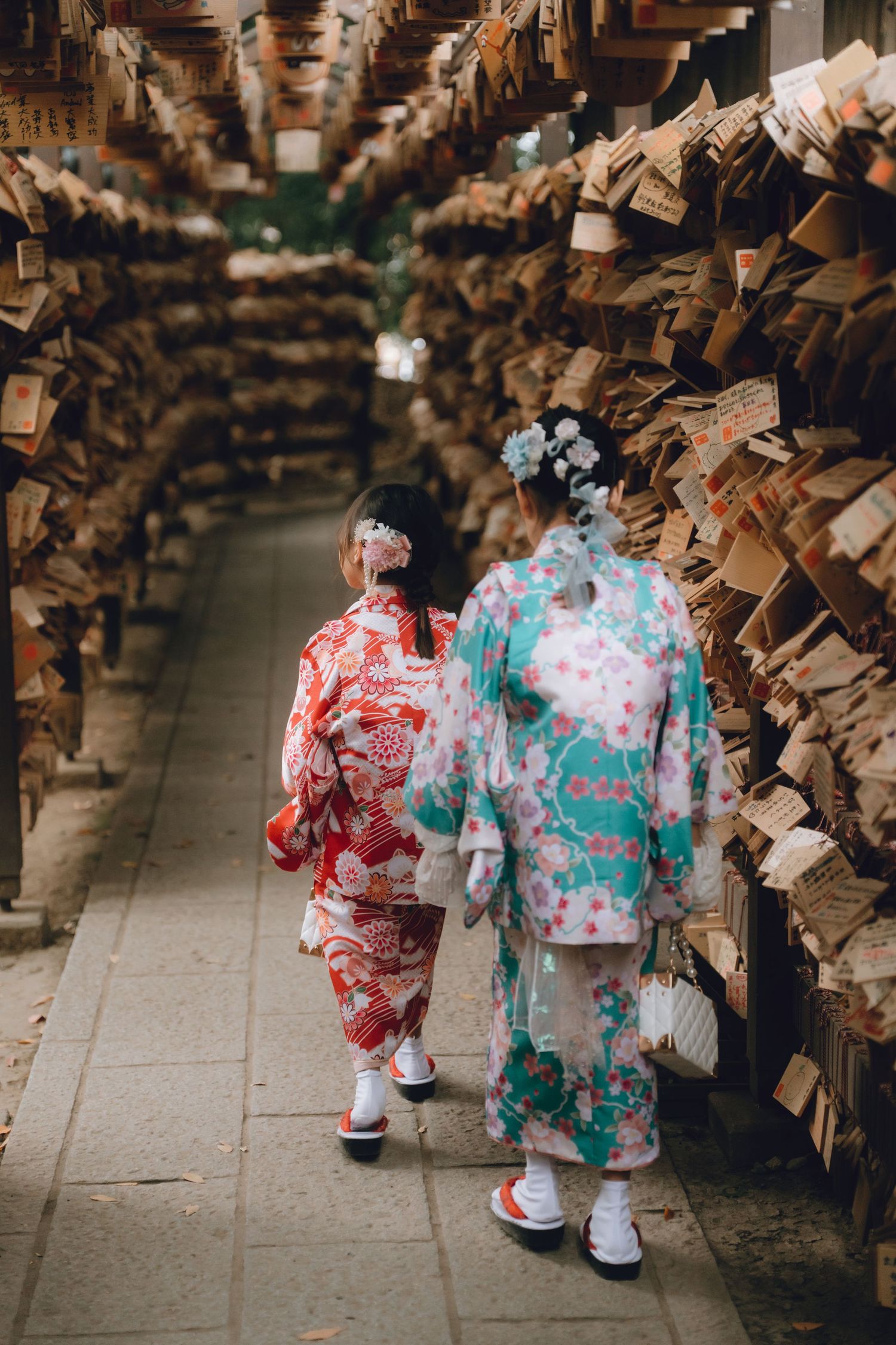 Two women in colorful traditional Japanese kimonos walk through a narrow path lined with wooden ema plaques at Hikawa shrine, Kawagoe, Japan