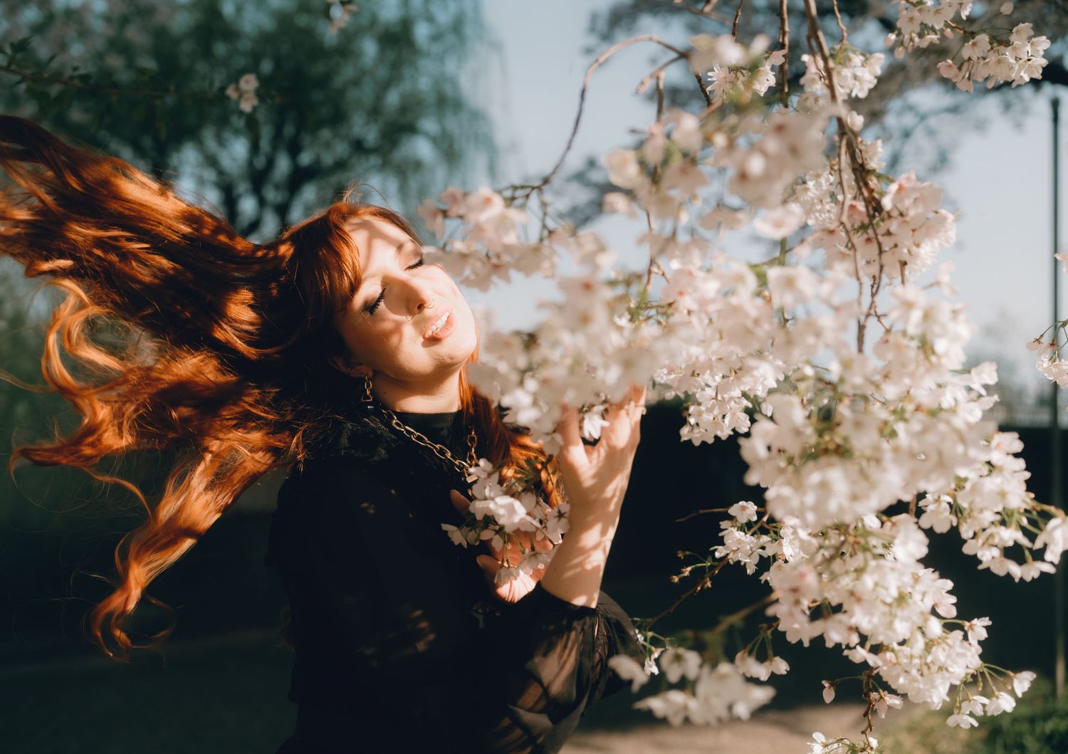 A red-haired woman with closed eyes poses near white cherry blossoms, her hair flowing in the breeze.