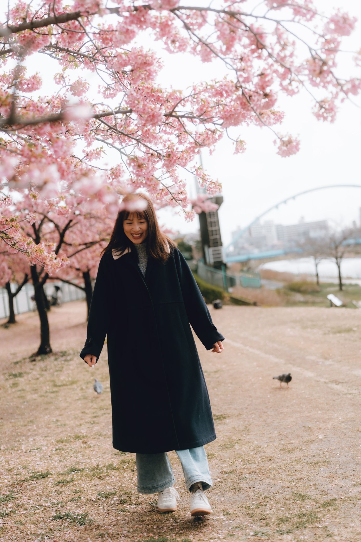 A smiling young woman in a navy coat walks under blooming pink cherry blossom trees in a scenic park.