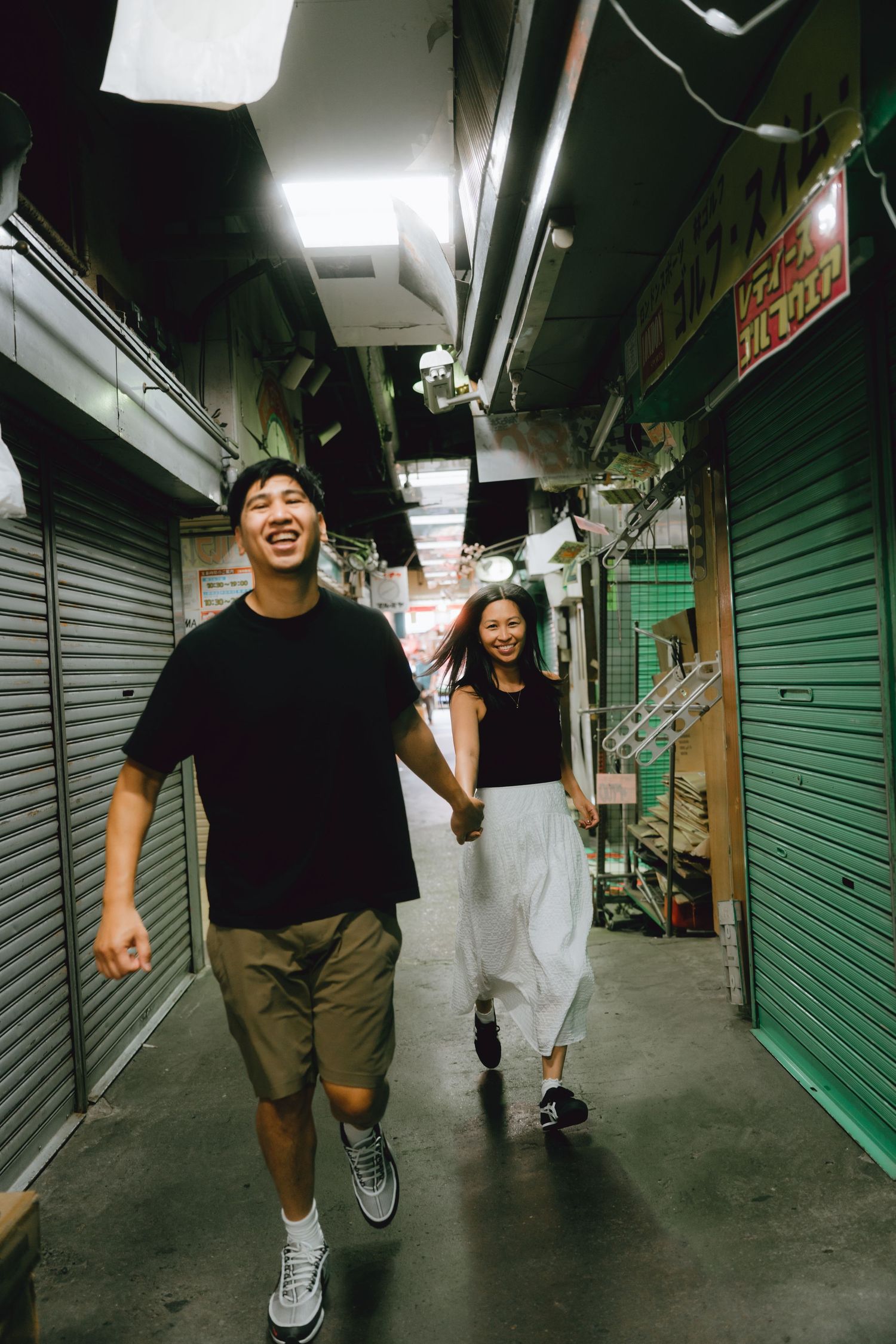 Happy Asian couple holding hands and running through a narrow urban alleyway at night, wearing casual black tops.