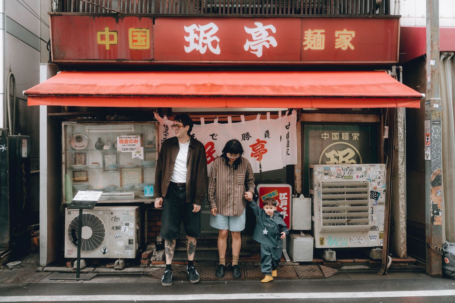 Three people standing outside a traditional Chinese noodle restaurant in Japan with red signage and storefront in Shimokitazawa, Tokyo.