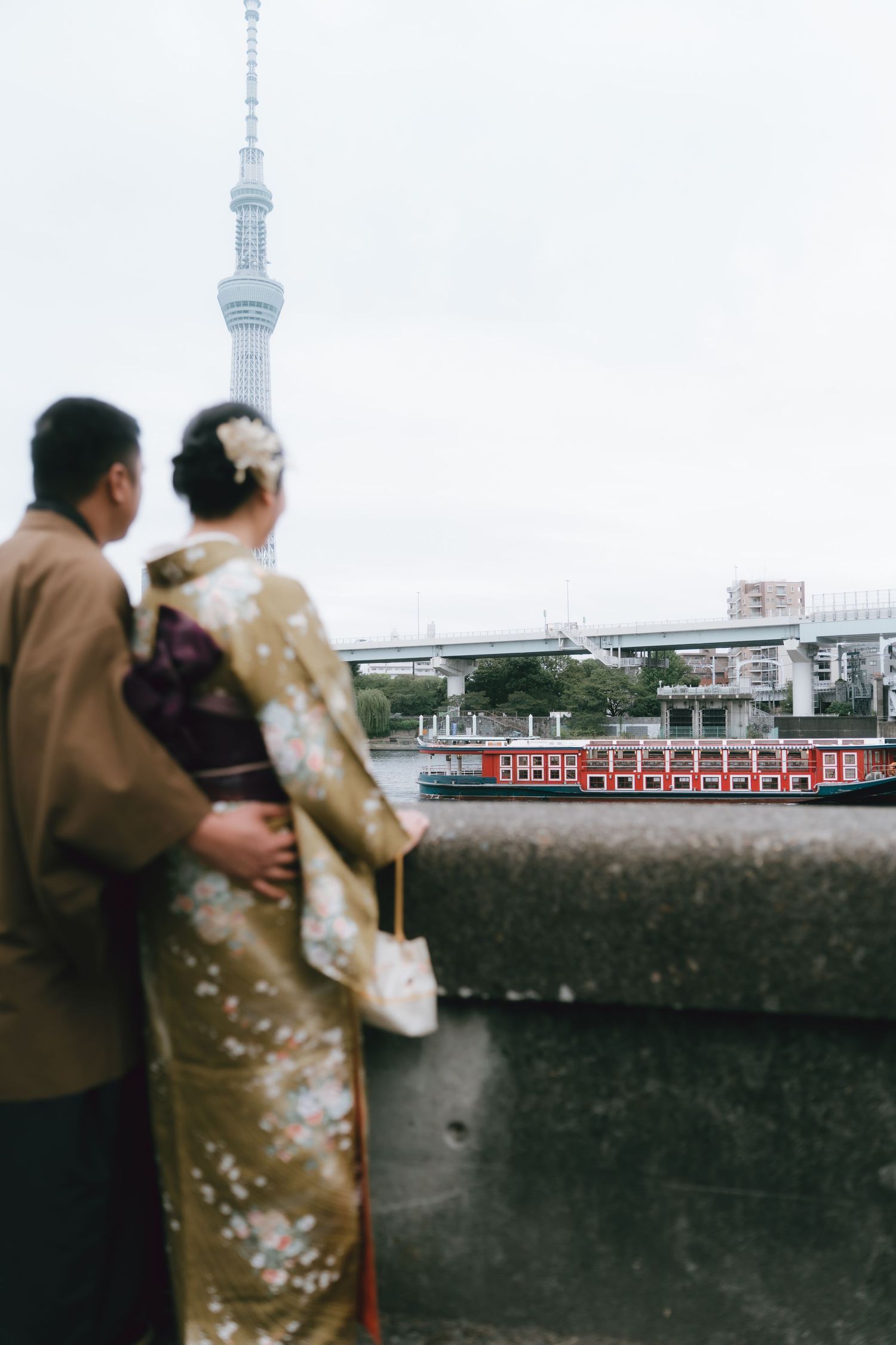 Couple in traditional Japanese kimono standing by Sumida river with Tokyo Skytree and red boat in background.