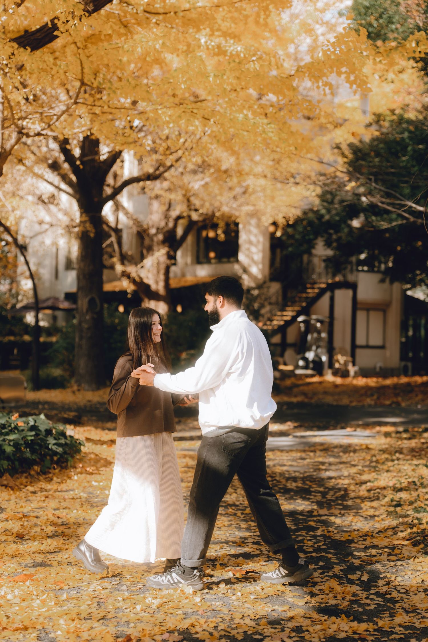 Couple dancing among golden ginkgo trees in autumn, surrounded by fallen yellow leaves and a charming building backdrop.