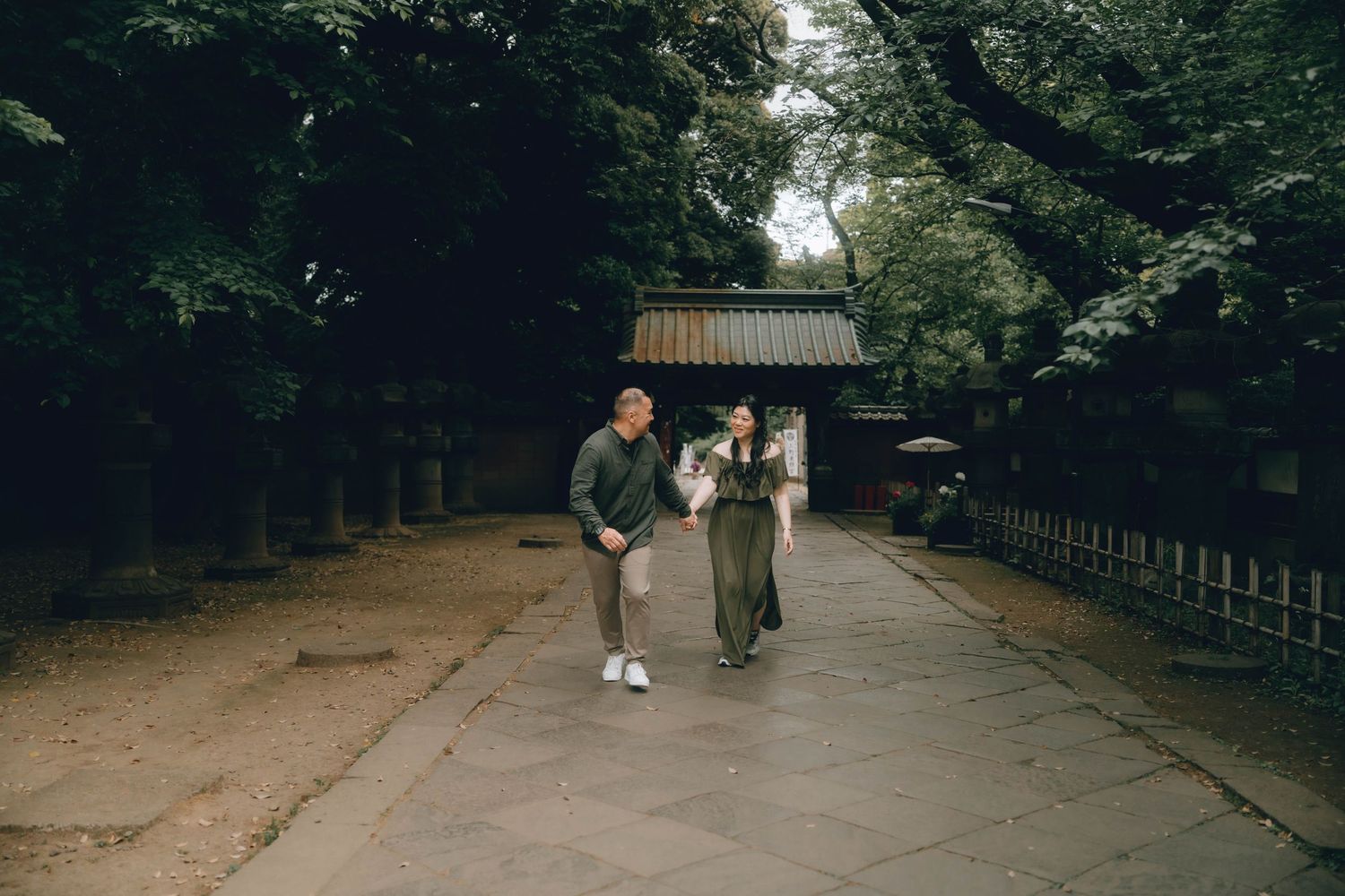 Couple walking along a stone path at a traditional Japanese shrine surrounded by lush green trees.