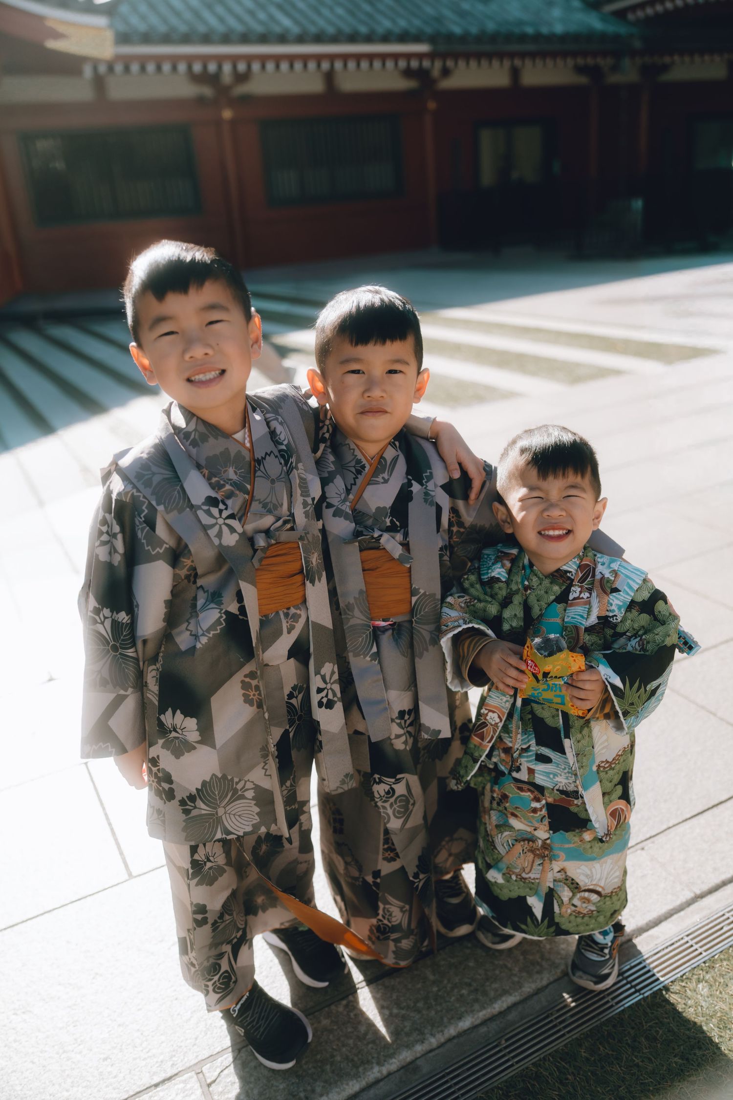 Three young boys wearing traditional Japanese kimono robes smile together outdoors in Asakusa.