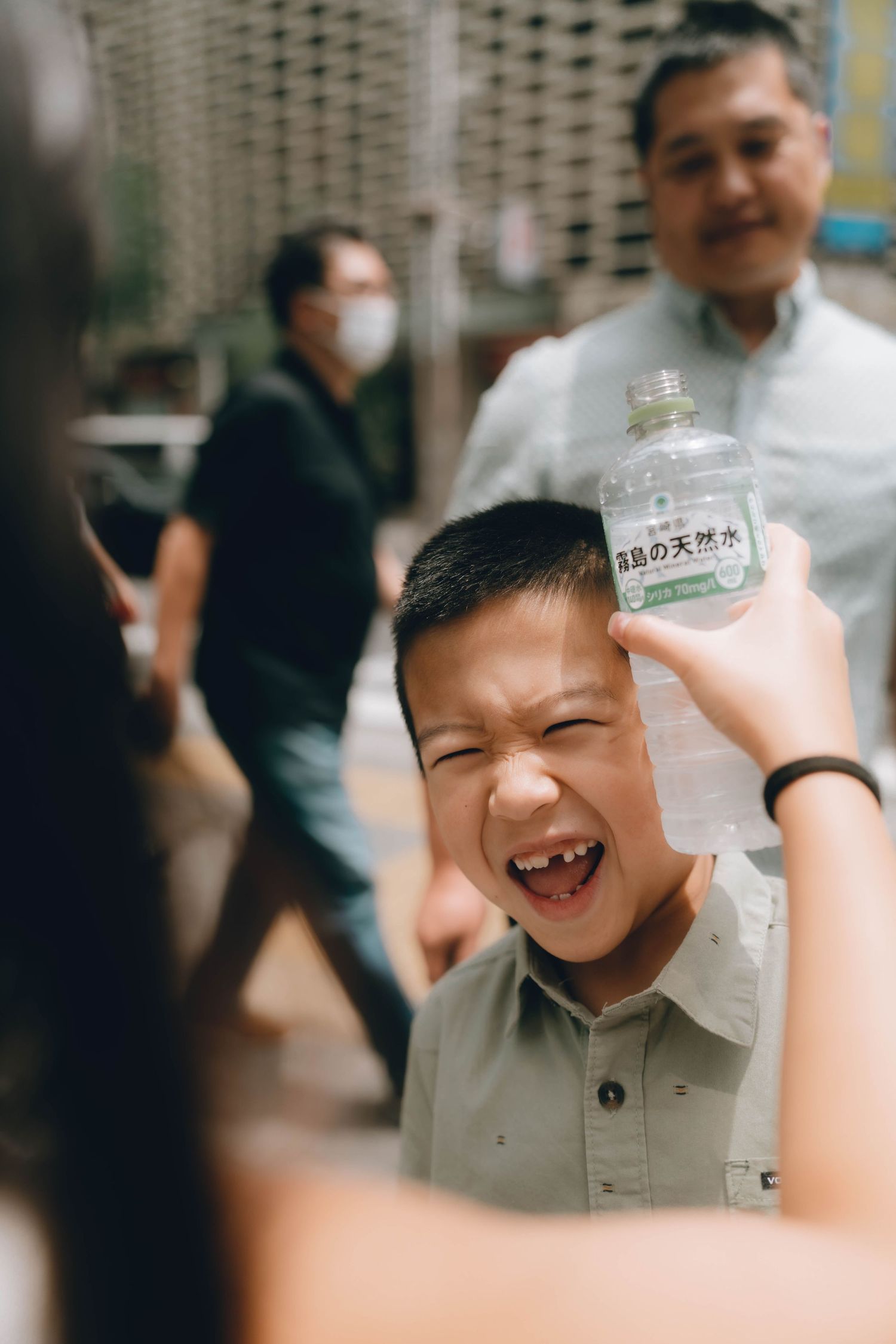 A young Asian boy makes a funny face while someone holds a Japanese water bottle near his head outdoors.
