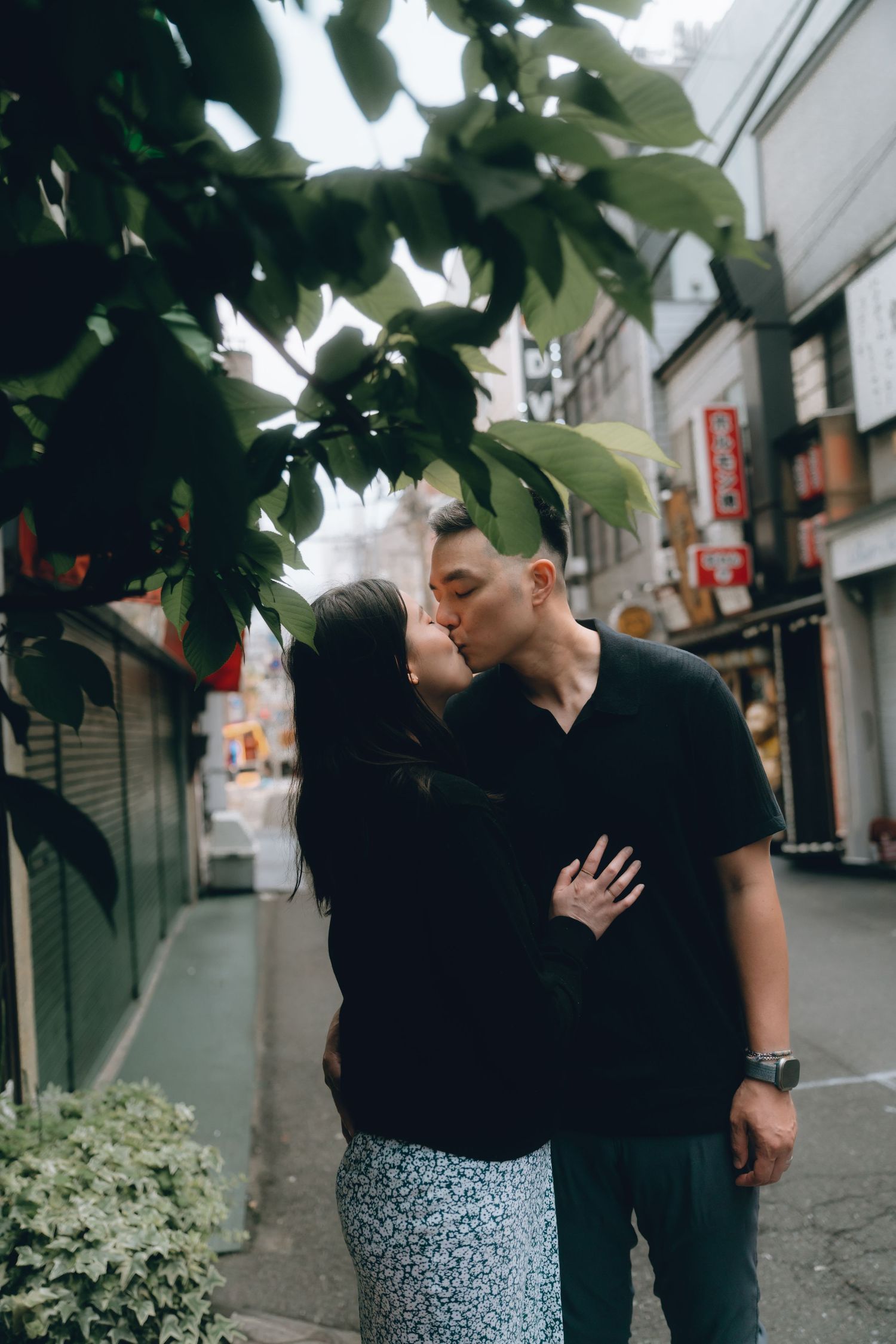 Couple sharing a romantic kiss on a quiet urban street in Japan, surrounded by lush green leaves and city storefronts.