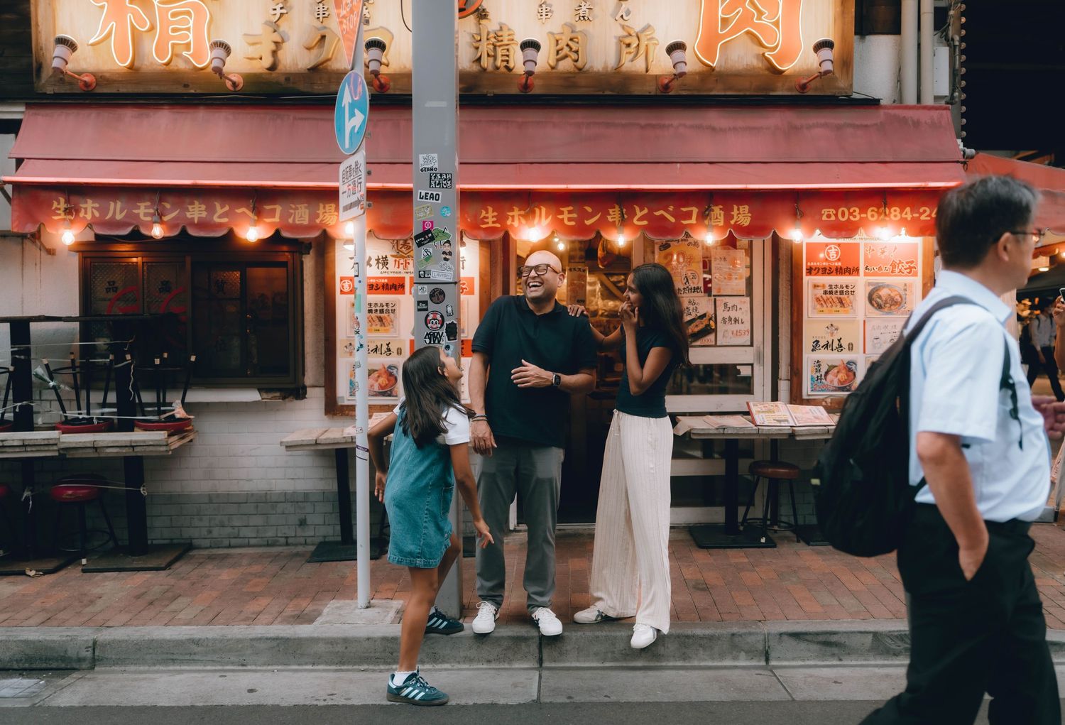 Family of three stands outside a vibrant Japanese restaurant at dusk, with neon signs and a passerby in foreground.