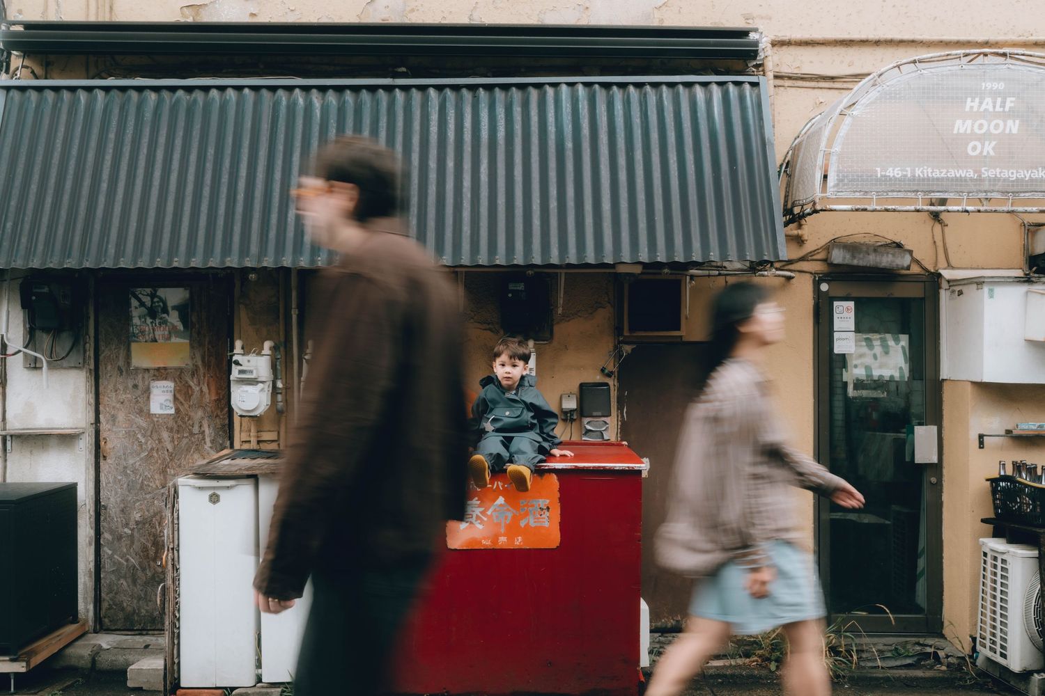 A child sits on a red cart outside a building while two blurred pedestrians walk past on a city street. Shimokitazawa, Tokyo.
