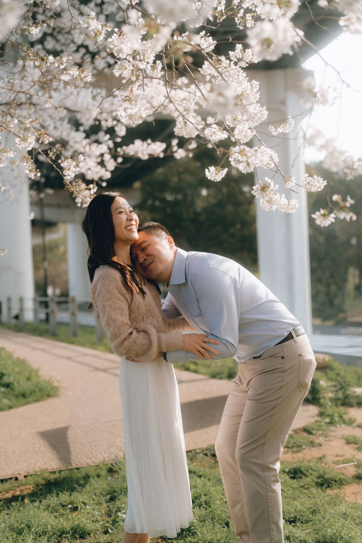 Couple embracing under blooming cherry blossom trees in spring, woman smiling joyfully in white skirt and beige sweater.