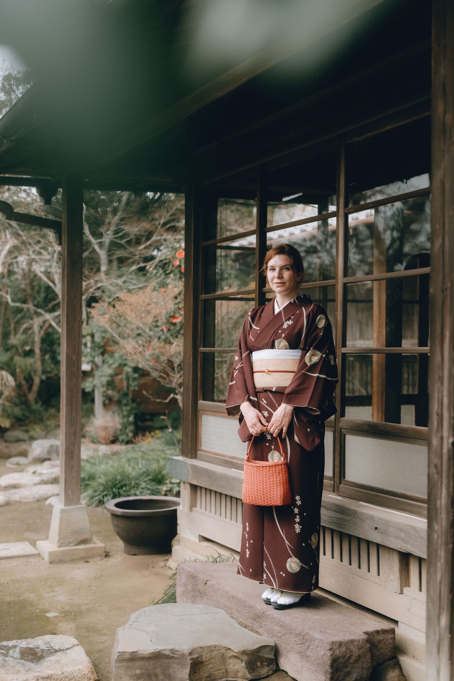 Woman in traditional brown Japanese kimono holding orange basket purse, standing on stone steps of a Japanese garden.Sawara, Chiba, Japan.
