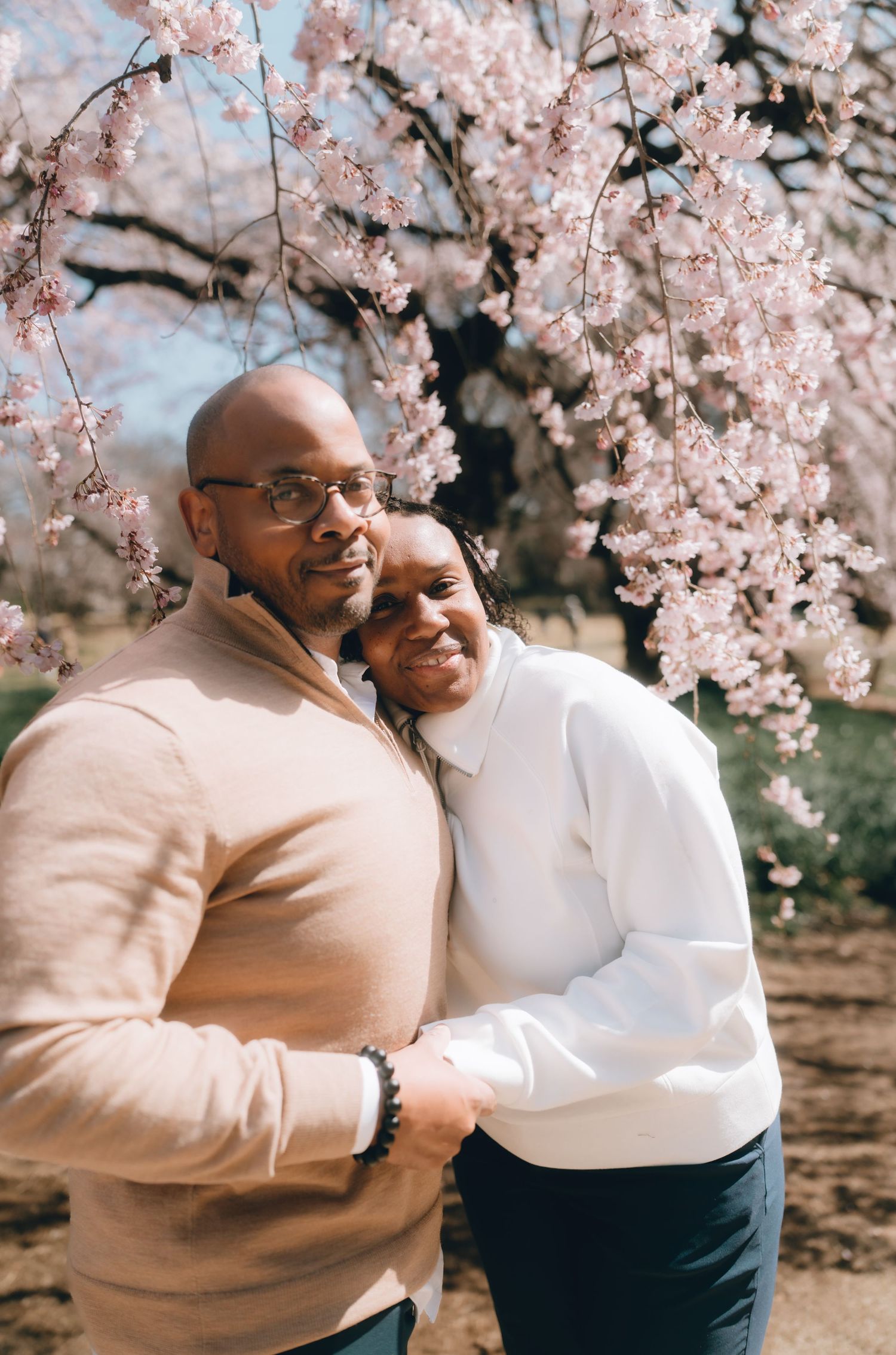 A couple embraces warmly in front of a blooming cherry blossom tree, smiling together in a serene outdoor setting.