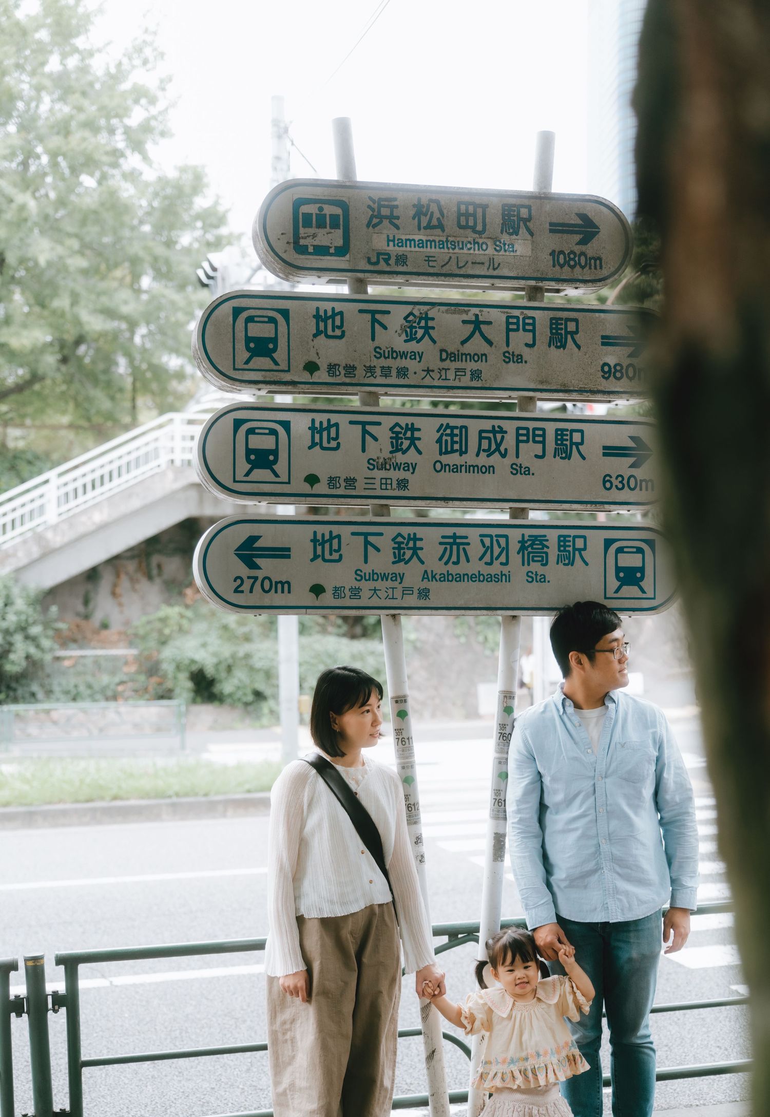 A family of three stands beneath Japanese subway directional signs in Tokyo, with a young child held between two adults near Tokyo tower.