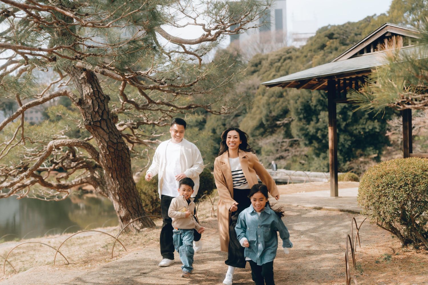 A happy family with two young children runs along a scenic garden path surrounded by trees and a traditional pavilion in Shinjyuku gyoen