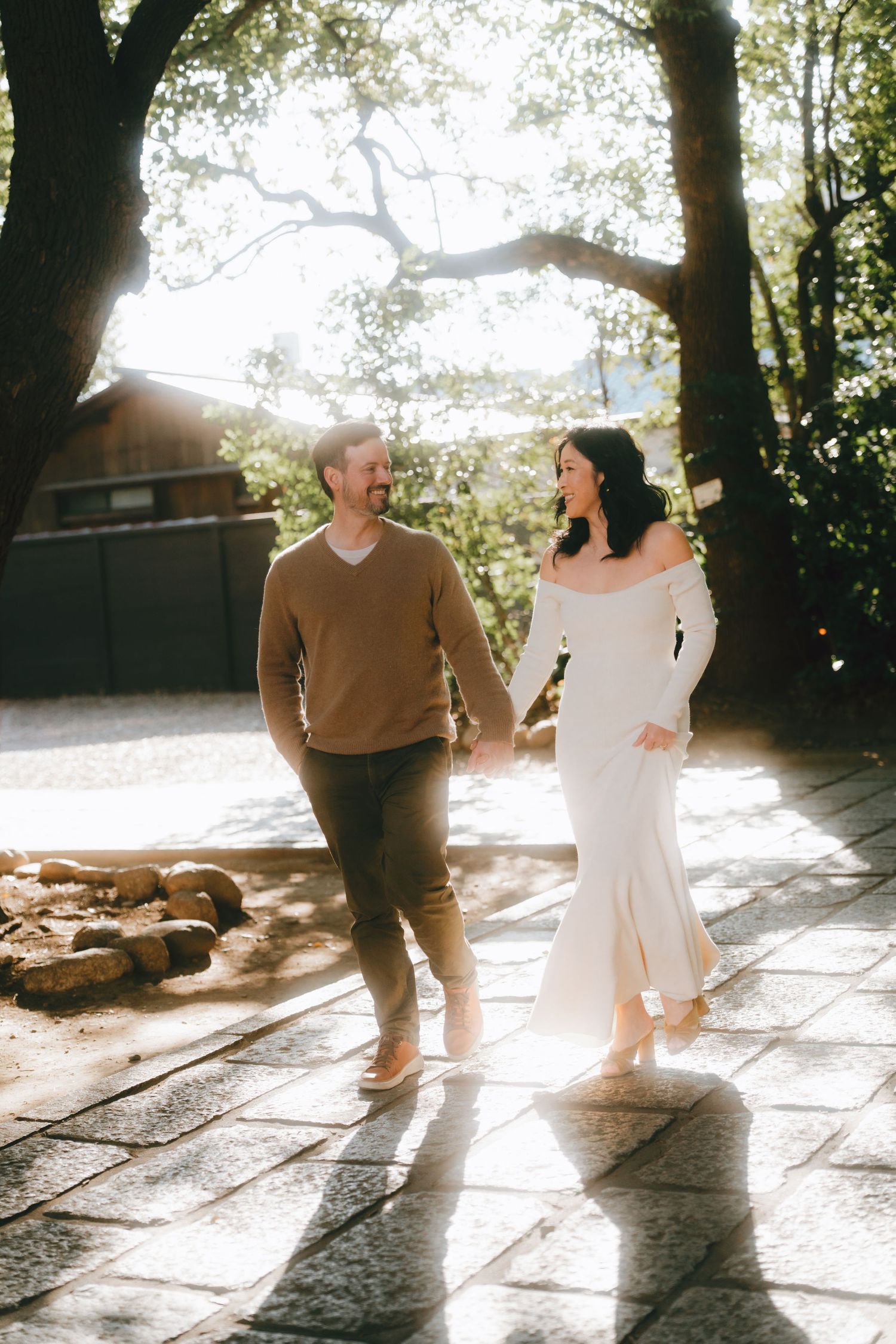 Couple holding hands walking on stone path in golden sunlight, woman in white dress, man in tan sweater, trees overhead.