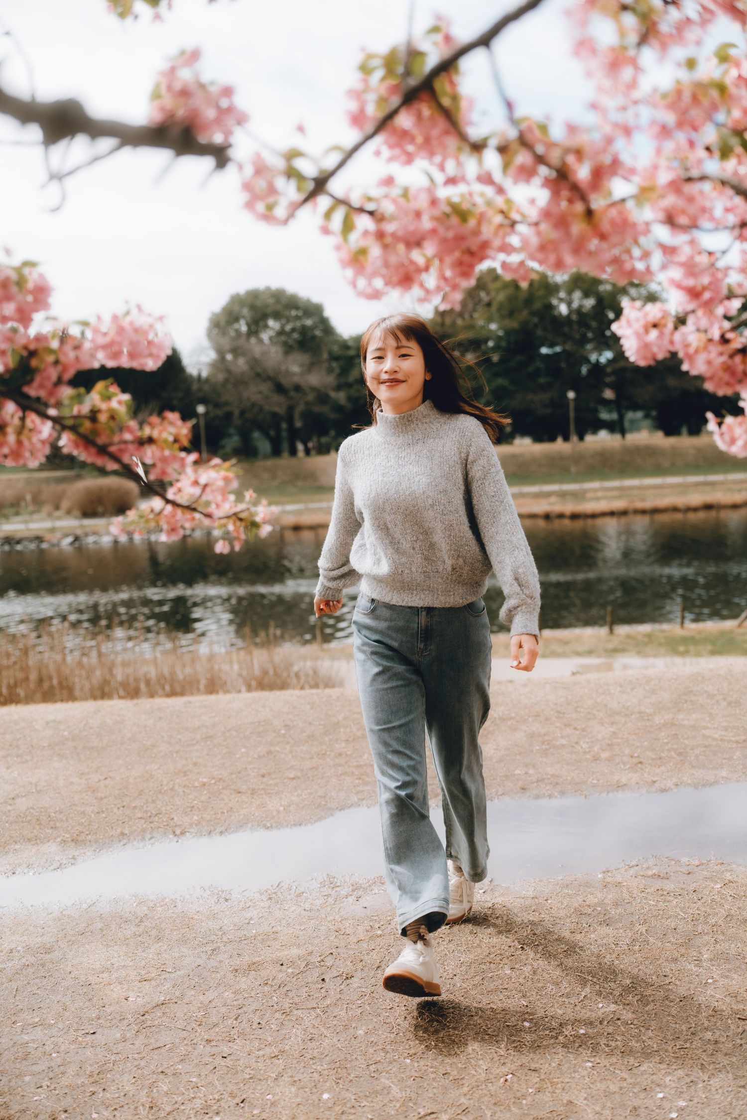 Young woman in gray sweater and jeans running near a cherry blossom tree by a pond in spring.