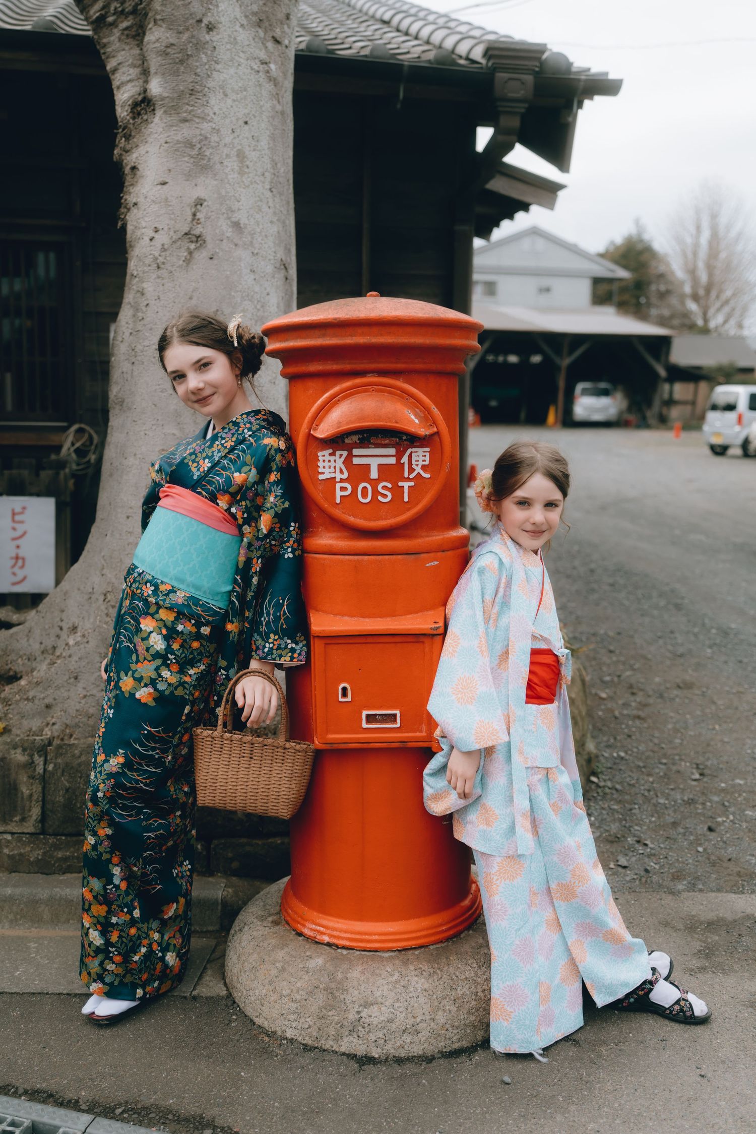 Two girls in traditional Japanese kimonos pose beside a red cylindrical post box sawara, cHiba, Japan.