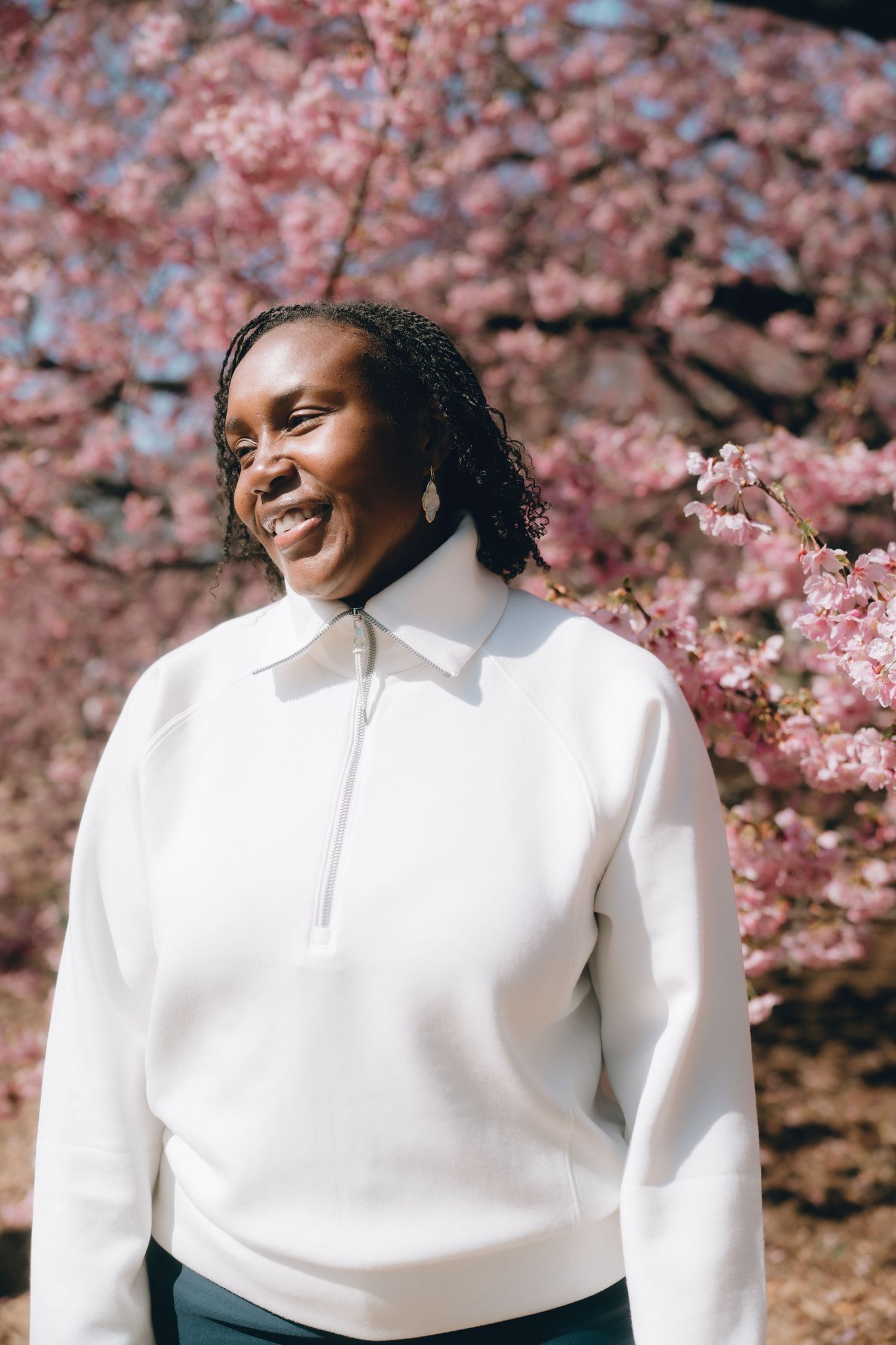Smiling Black woman in white zip-up sweatshirt standing outdoors in front of pink cherry blossom trees.
