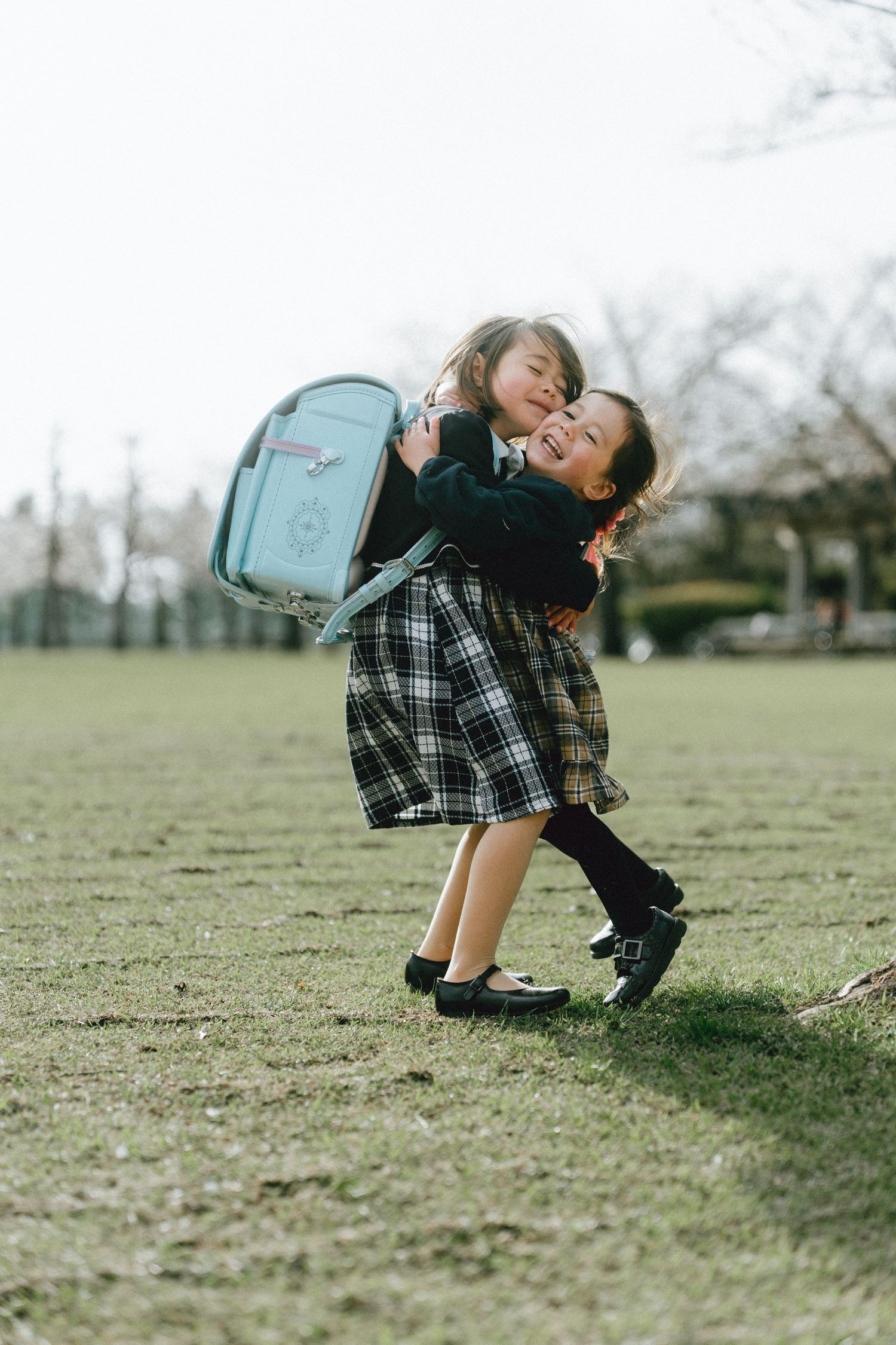 Two young girls in plaid uniforms share a joyful hug outdoors, one wearing a light blue randoseru backpack.