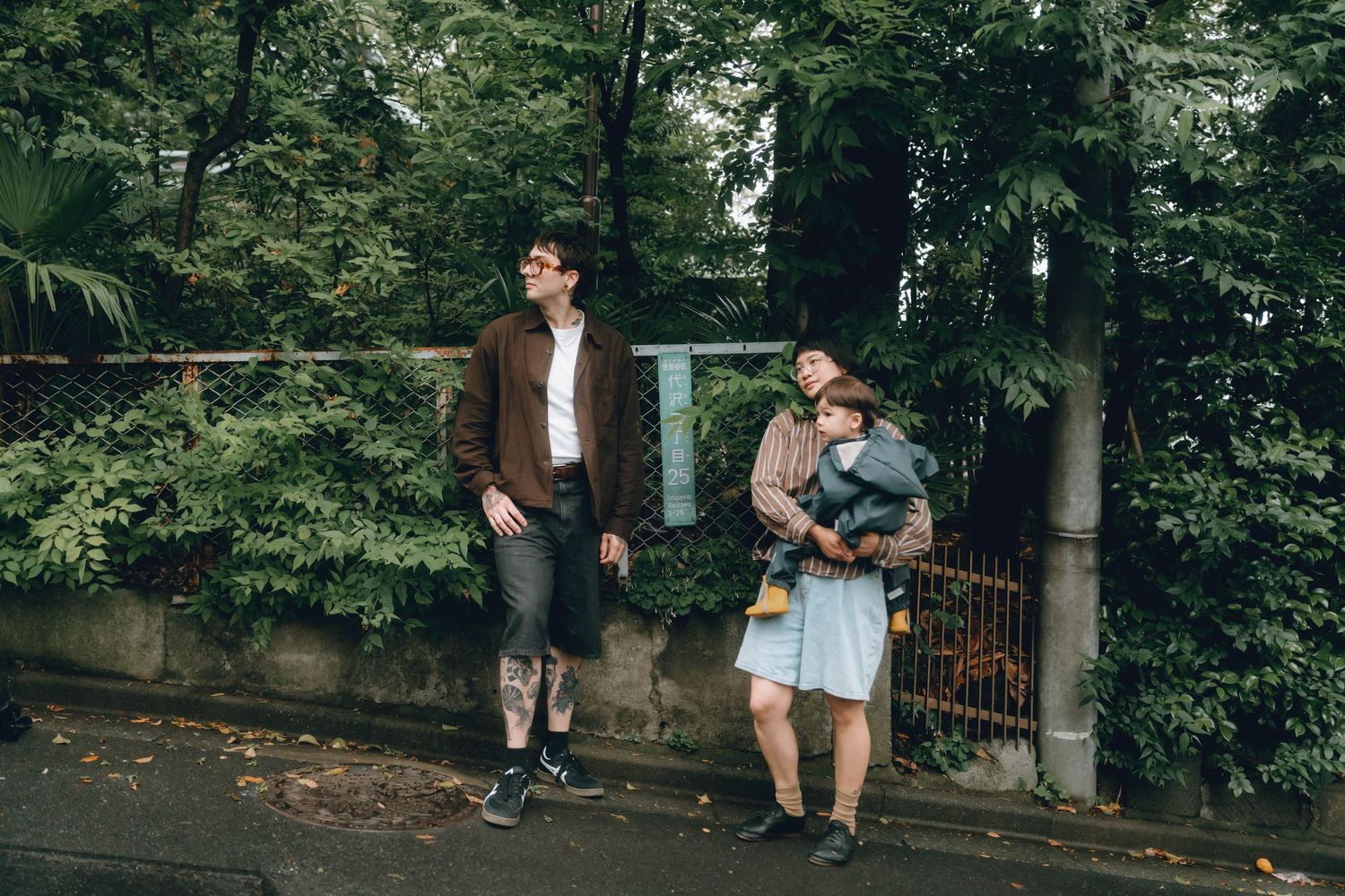 Two people standing on a wet sidewalk surrounded by lush green trees and bushes on a cloudy day in Shimokitazawa.