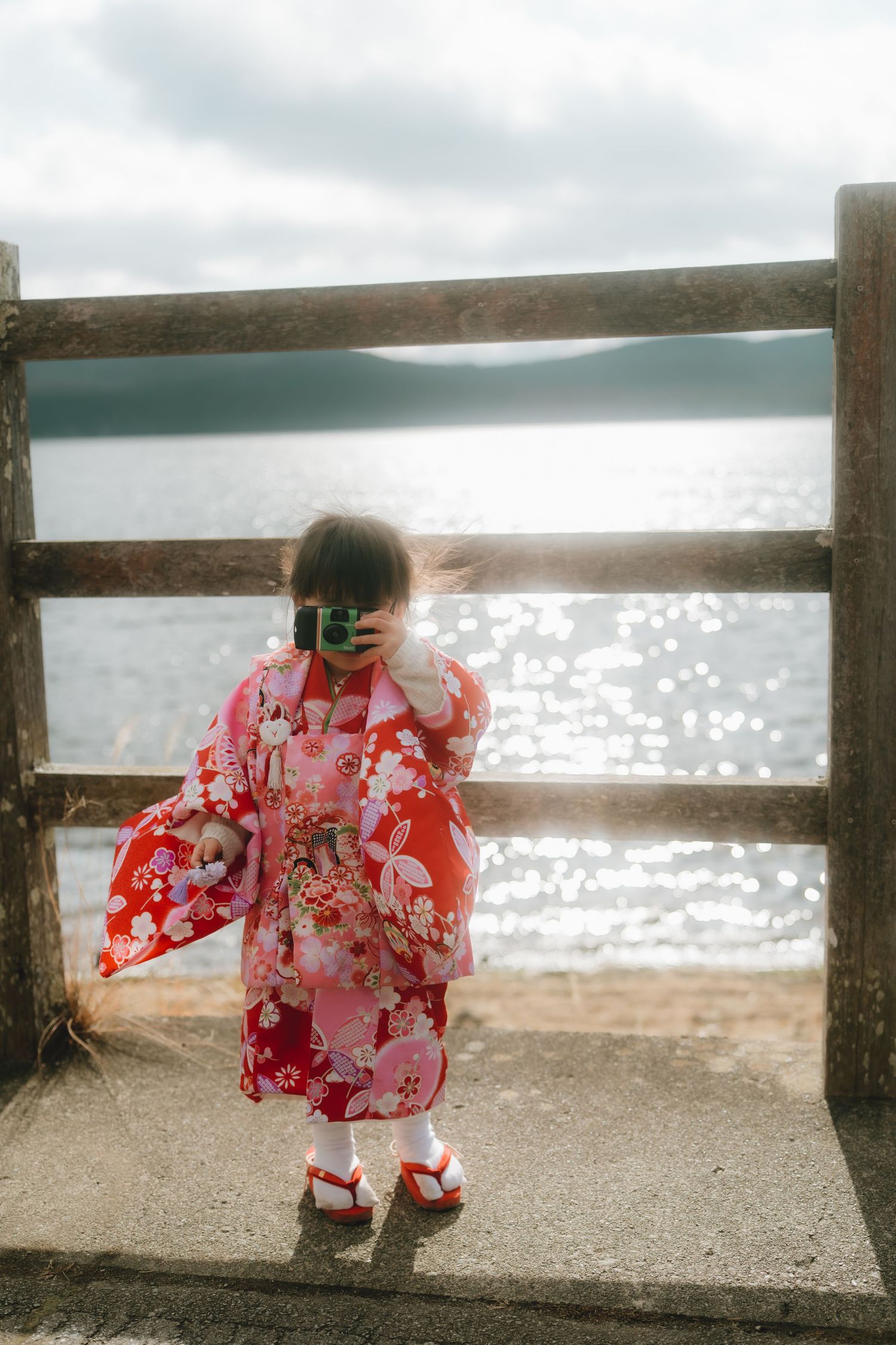Young girl in red floral kimono photographing lake yamanaka from a wooden railing