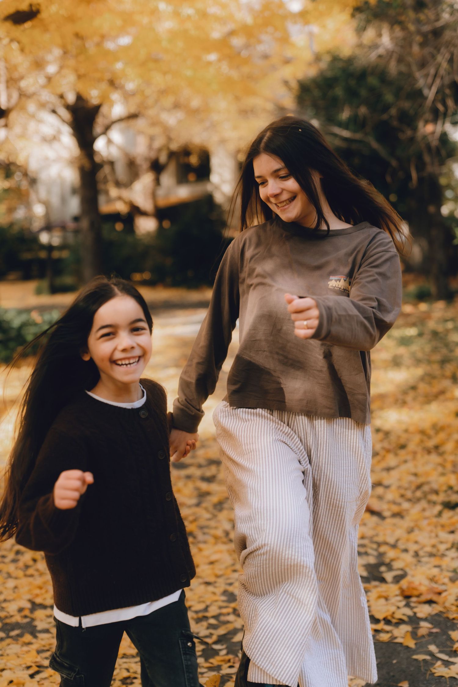 Two smiling girls walk hand-in-hand through a park surrounded by golden autumn leaves in Hibiya park, Tokyo.