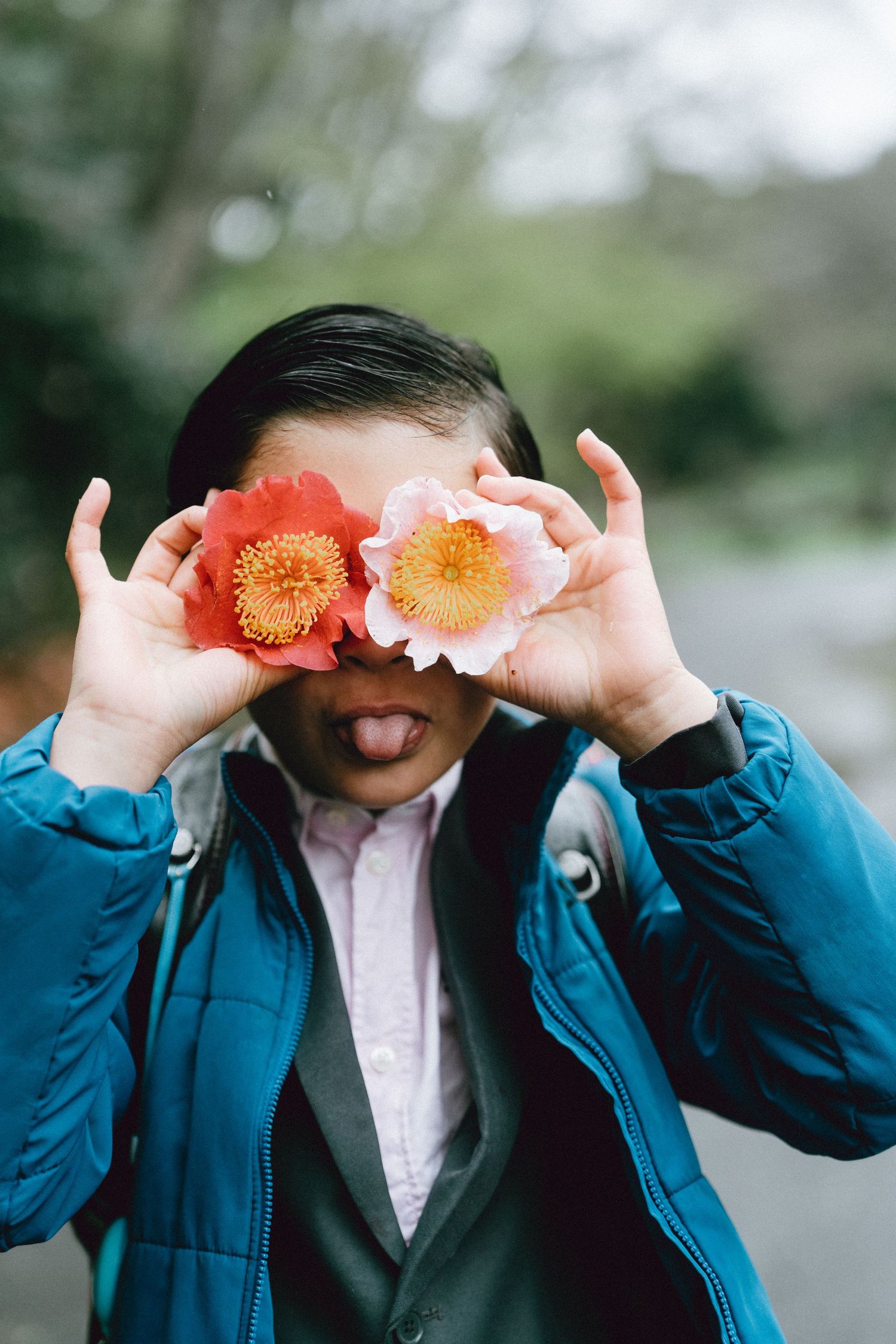 A playful person holds two pink camellia flowers over their eyes while sticking out their tongue, wearing a teal jacket.