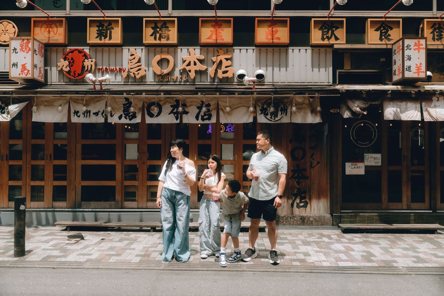 Family of four posing together on a street in front of a traditional Japanese restaurant with lanterns and kanji signage in Shinbashi, Tokyo
