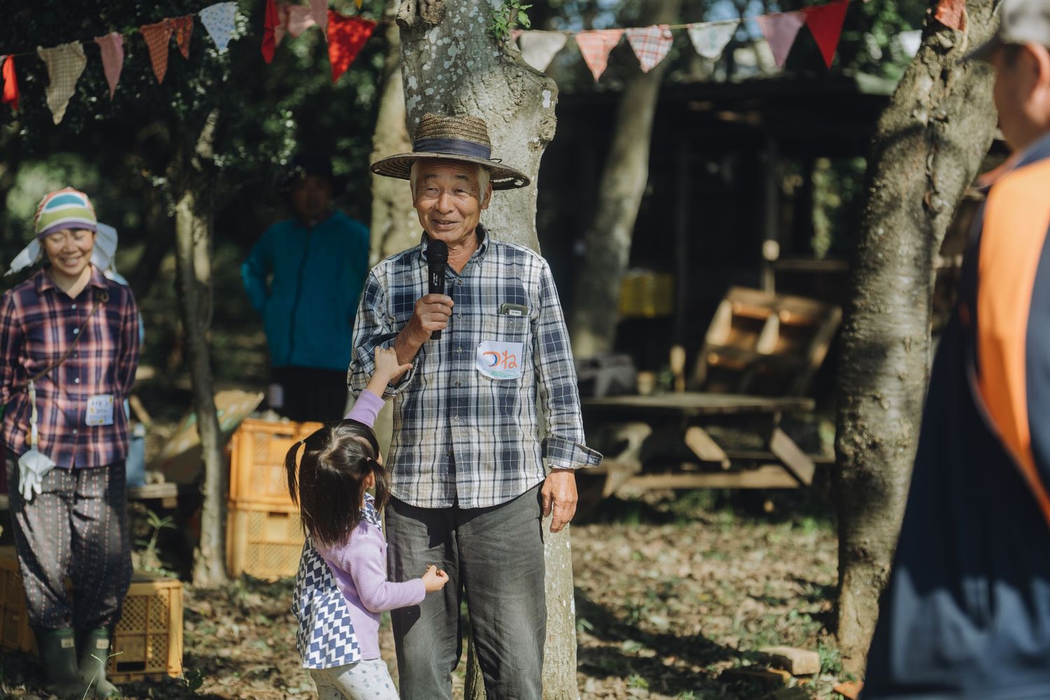 Elderly man in plaid shirt and straw hat smiles at a child during an outdoor festival with colorful bunting.