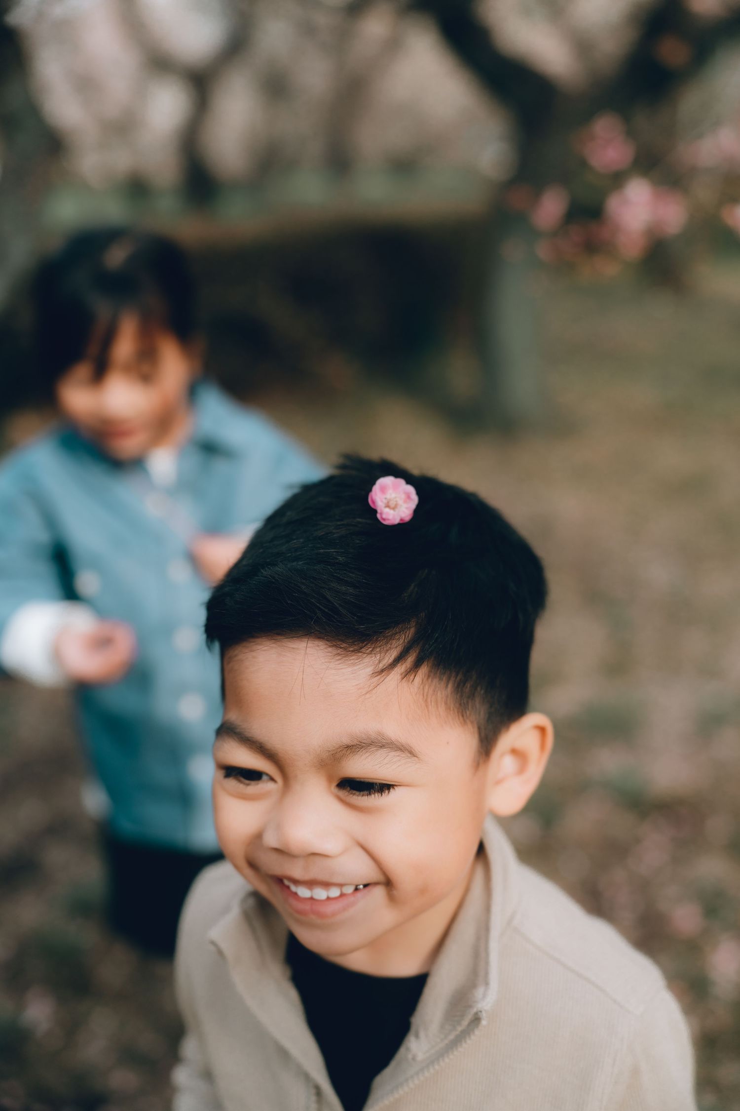 Smiling Asian boy with pink flower in hair outdoors, girl in blue jacket blurred in background near blooming trees.