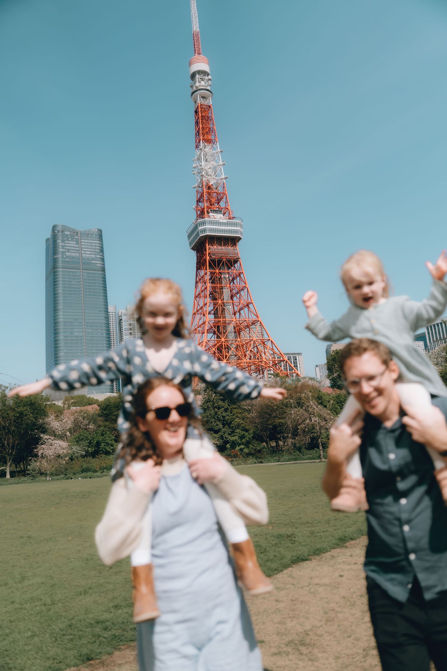 A happy family with two young children on their shoulders poses in front of Tokyo Tower on a sunny day.