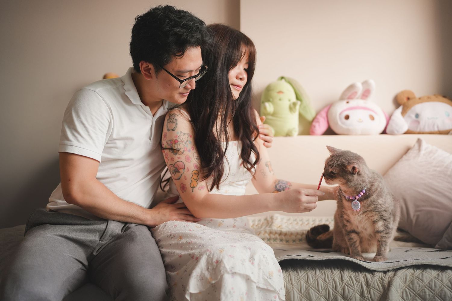 Asian couple sitting on bed with stuffed animals, interacting with a small kitten between them.