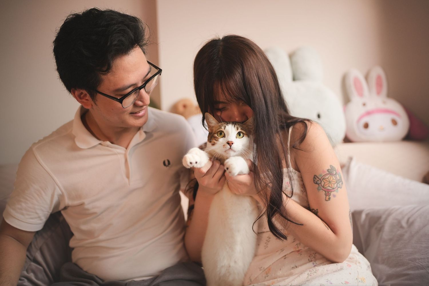 Asian couple on bed, woman holding small white cat up close to her face while man smiles.