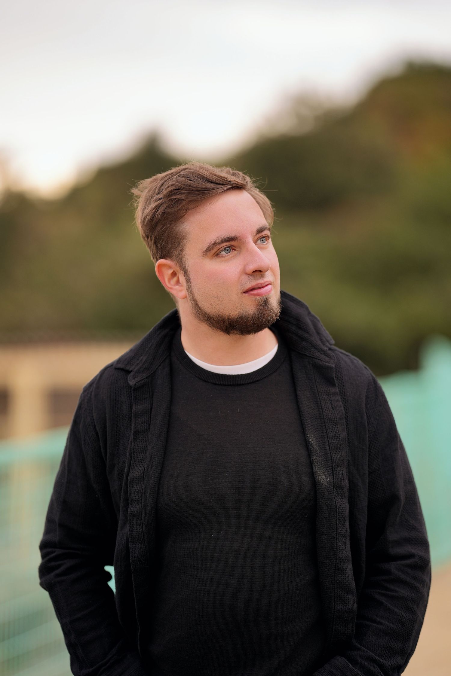 Young man with beard wearing black jacket standing outdoors with blurred green trees in background.