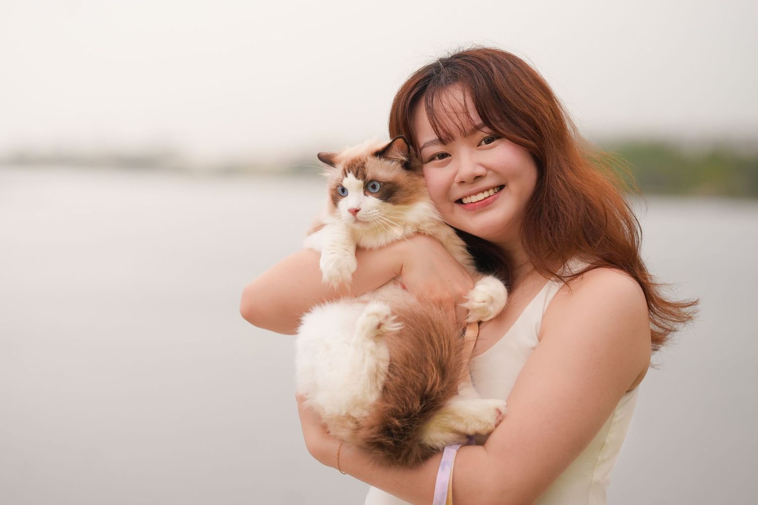 Smiling young woman holding a fluffy Ragdoll cat outdoors near a lake.