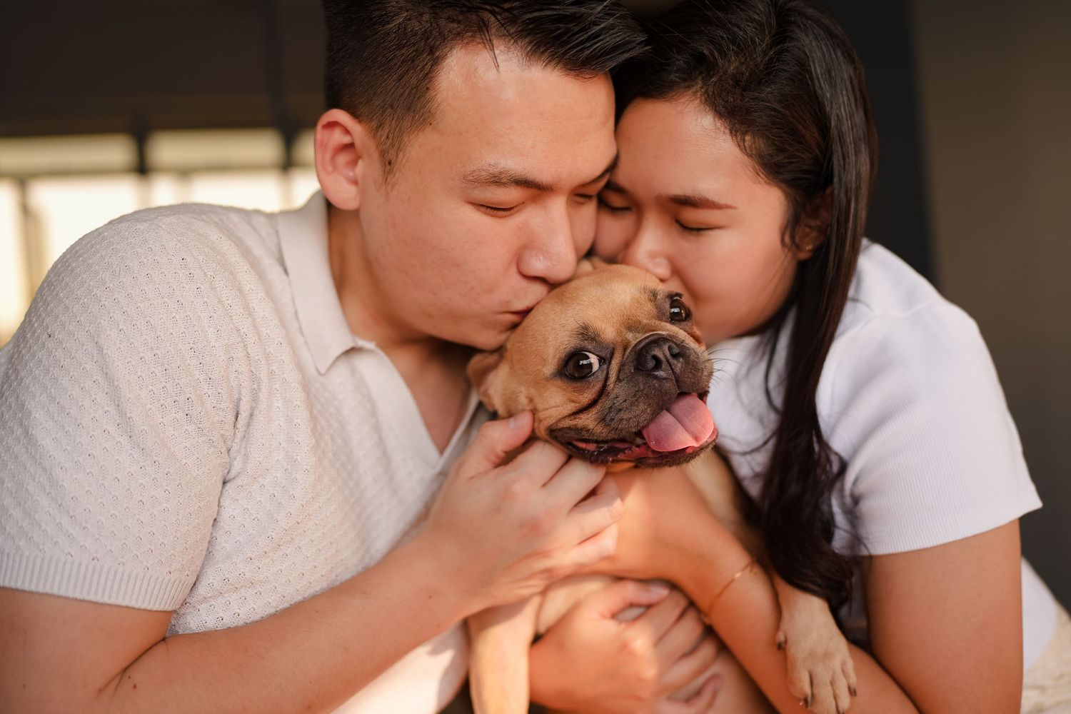 A couple kisses their adorable French Bulldog puppy together indoors.