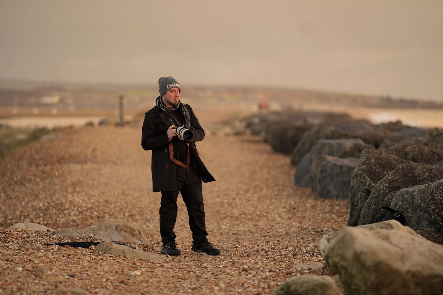 Man standing on rocky beach in winter coat and grey hat holding camera on overcast day.
