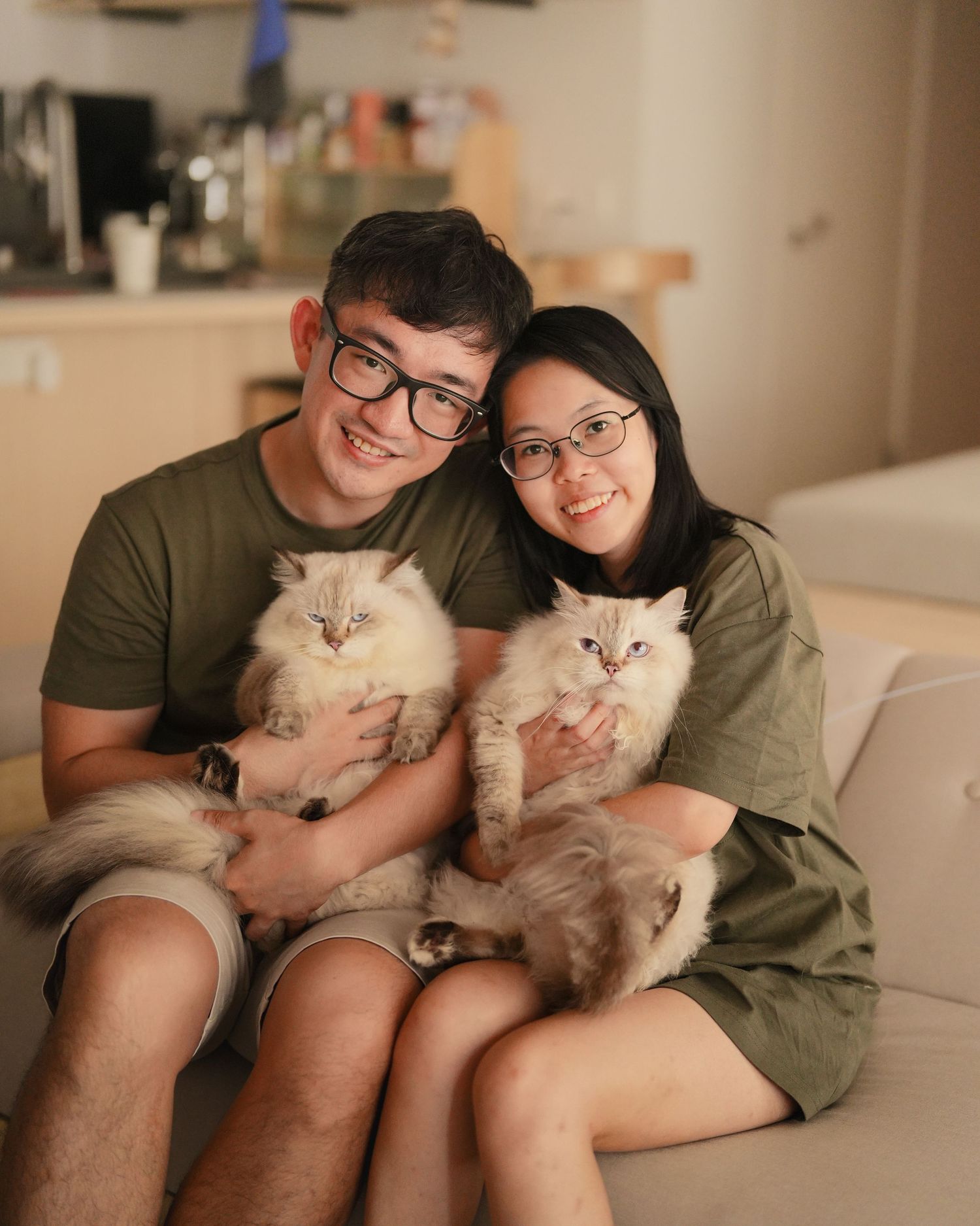 A smiling couple wearing glasses holds two fluffy Ragdoll cats while sitting on a white sofa.