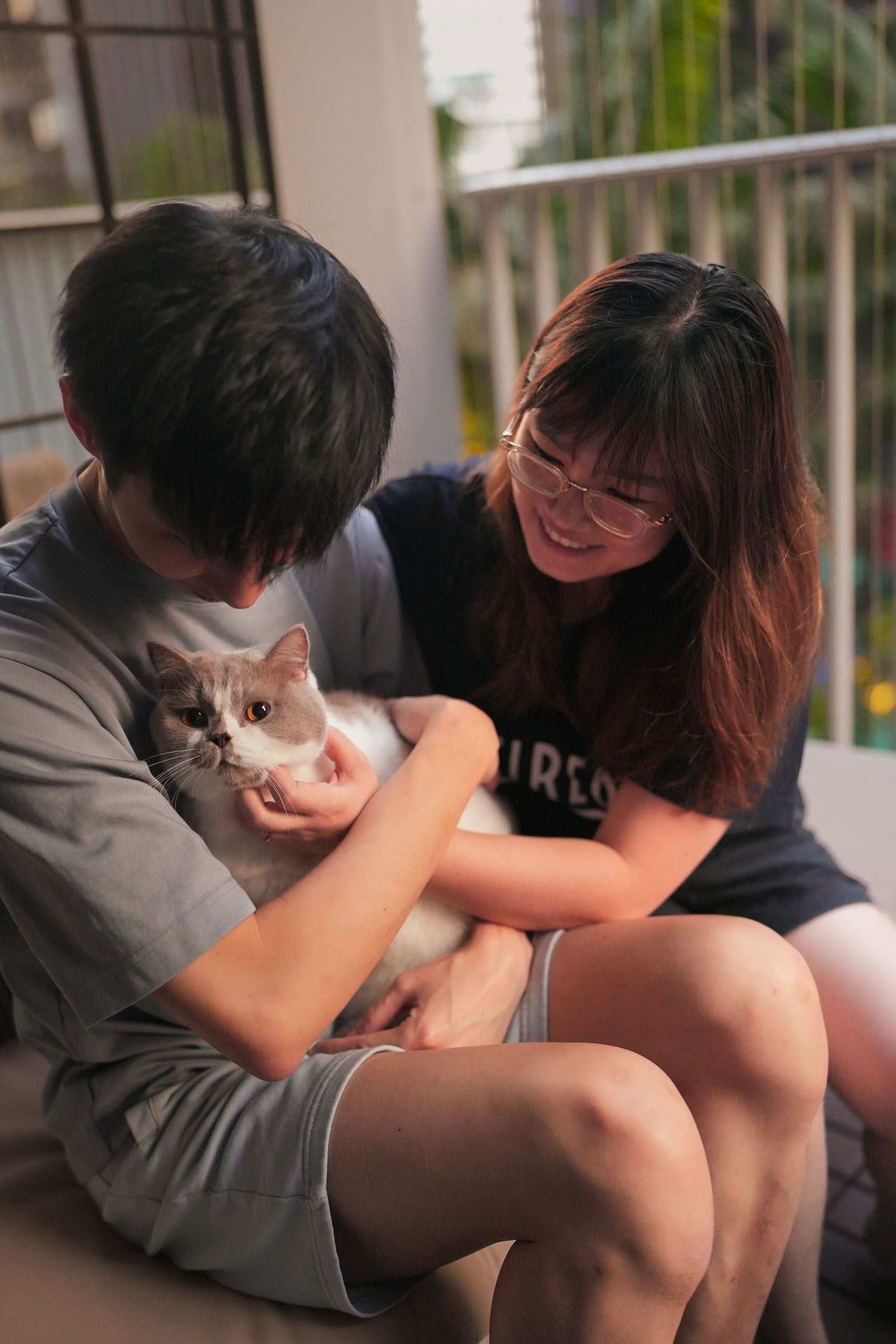 Young Asian couple sitting on balcony floor, smiling while petting a white cat together.