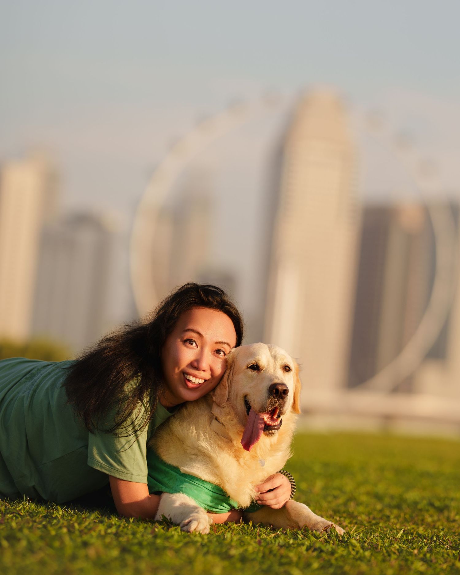 Smiling woman lying on grass hugging a Golden Retriever with city skyline behind.