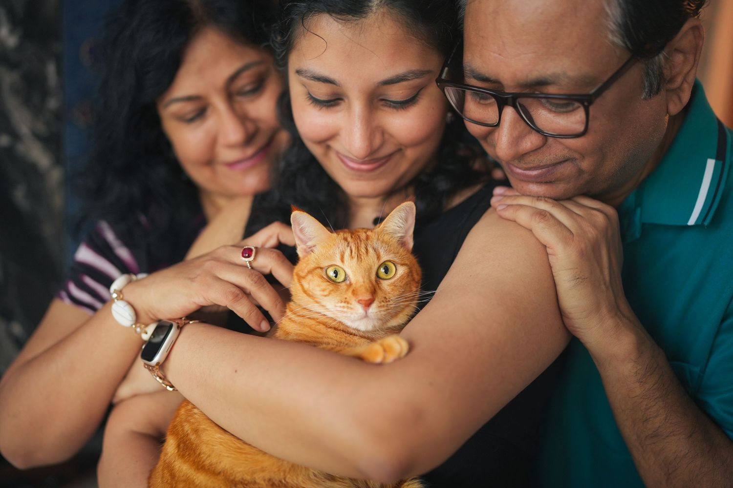 Happy family of three closely bonding with an orange tabby cat, smiling together indoors.